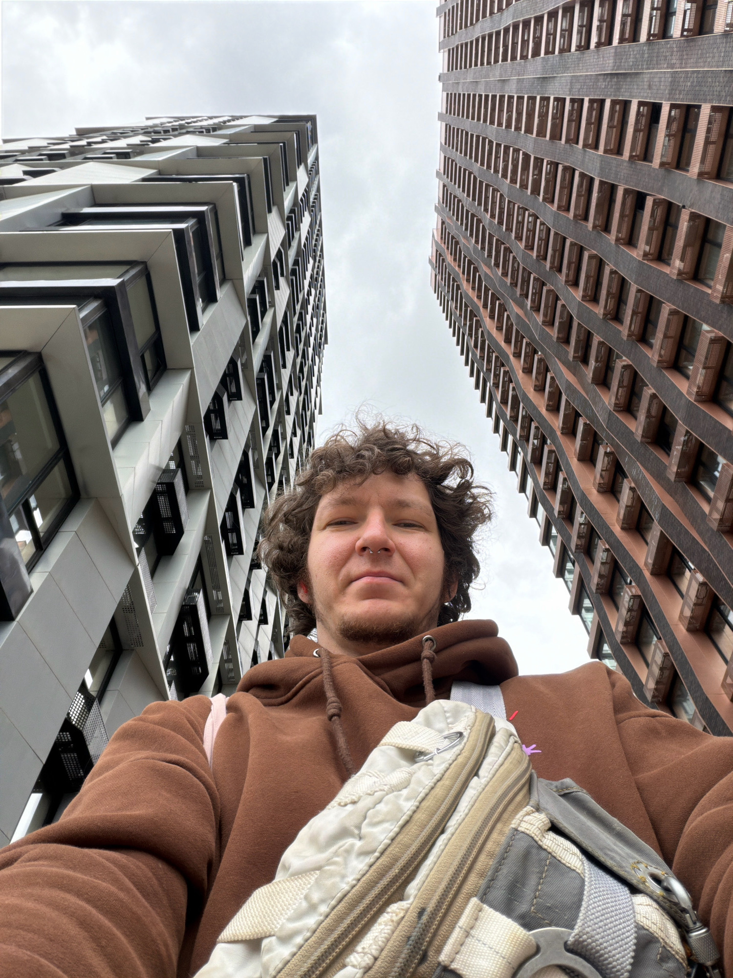 A low-angle, wide shot captures a person wearing a brown hoodie and a cross-body bag, standing between two modern high-rise buildings. The building on the left features a white facade with a geometric, angular design and dark windows. The building on the right is characterized by a brown brick facade with rows of windows. The sky is overcast. The image appears to be taken in Moscow, Russia.