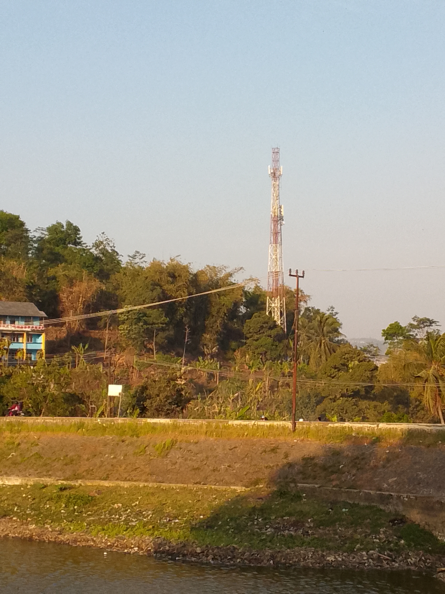 The image depicts a serene landscape on the outskirts of Padalarang, Indonesia, bathed in the warm light of what appears to be late afternoon. Dominating the skyline is a prominent telecommunications tower, a lattice structure painted in alternating bands of white and red, signifying its height and purpose. Below the tower, a lush green hillside is densely populated with a variety of trees and vegetation, characteristic of the region's tropical climate.
On the left side of the frame, a multi-story building with a blue and yellow facade stands as a testament to local architecture, its windows suggesting residential or commercial use. A single motorcycle is visible on a paved road that skirts the base of the hill, indicating vehicular access to the area. The road is lined with low-lying greenery and a retaining wall, separating it from the water body in the foreground.
The foreground is occupied by a calm body of water, its surface reflecting the muted colors of the sky and surrounding foliage. The bank of the water is a mix of rough terrain, with patches of grass and scattered debris, hinting at the natural, less manicured environment. A lone utility pole with a few crossbars stands near the telecommunications tower, a mundane but essential piece of infrastructure against the backdrop of nature. The overall impression is one of a tranquil, semi-urban setting where natural beauty meets functional development.