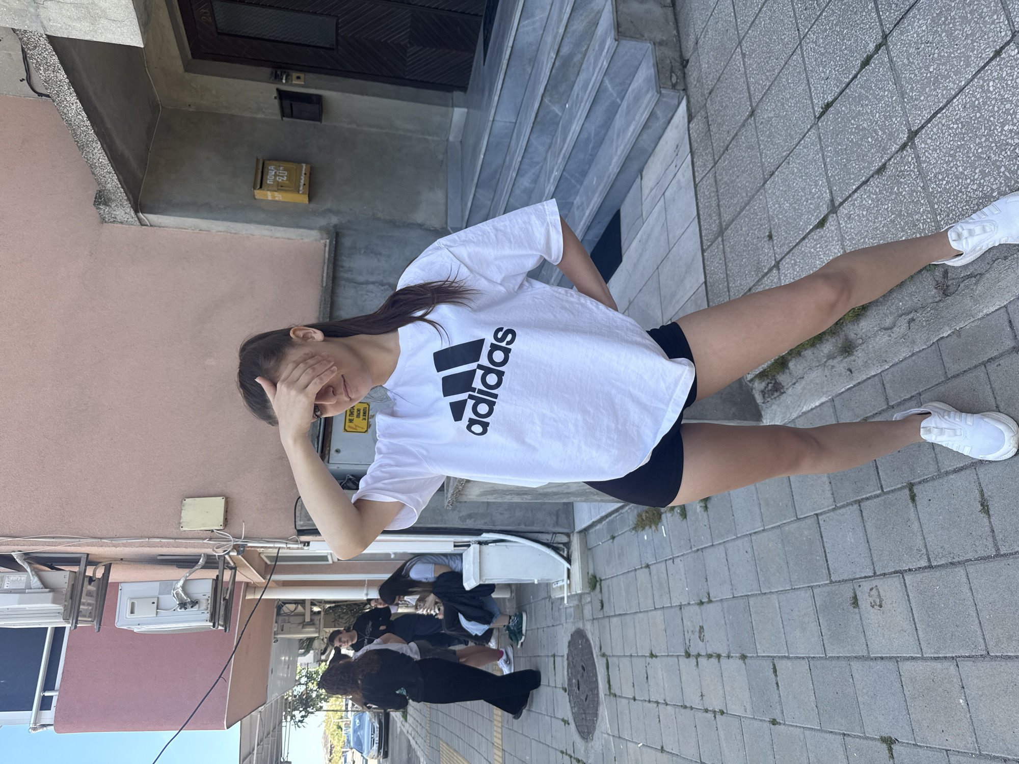 A young woman wearing a white Adidas t-shirt and black shorts stands on a paved sidewalk in Pomorie, Bulgaria. Her right hand is raised, obscuring part of her face. She is wearing white sneakers. Behind her, a group of people is gathered, with cars visible in the street. The architecture includes buildings with pink facades and concrete stairs leading to a dark-colored door. An electrical box and air conditioning units are attached to the building's exterior. A yellow mailbox is also visible on the wall.