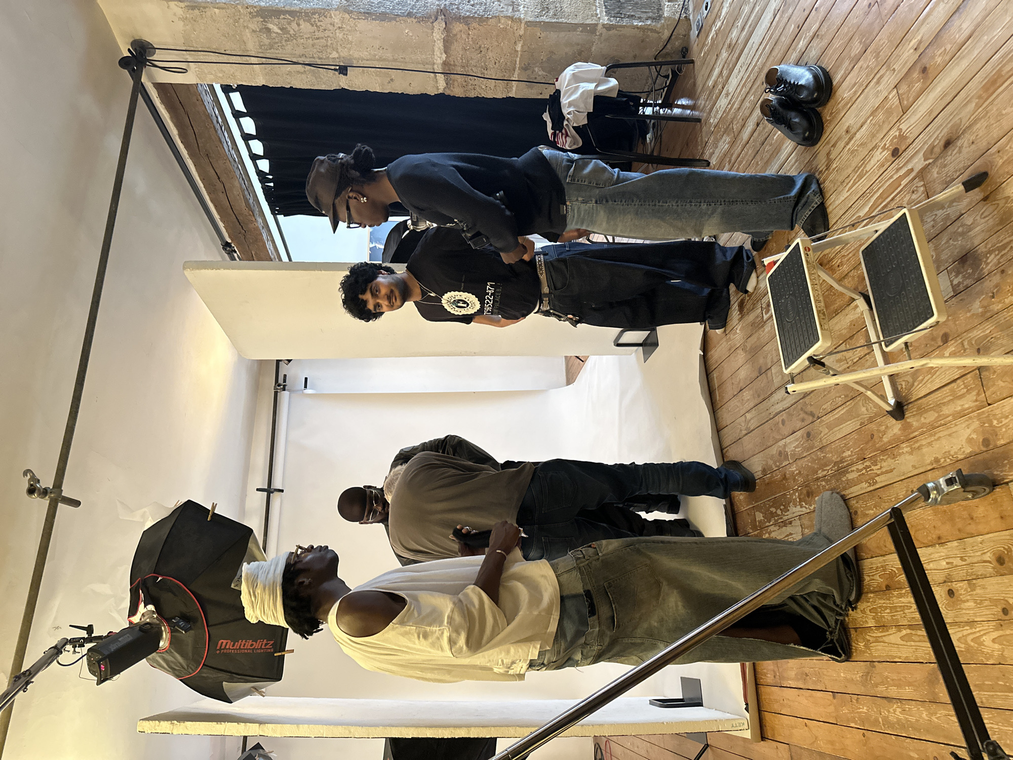 The image depicts a behind-the-scenes scene of a fashion photoshoot in what appears to be a studio in Paris, France. There are five people visible in the image. Three people are standing in the foreground, the middle person is turned so that his face is visible. The person on the right is standing next to him and looking down, with glasses, a brown hat and black clothing. There are two other people standing next to them. One is standing next to a white lighting screen, looking upwards, and the person next to him is wearing glasses and a light brown top. A "Multiblitz" lighting setup is positioned to the left.
The studio has a wooden floor and white walls, with exposed architectural elements suggesting an older building. Near the back of the image, there is a black curtain and a chair with clothes on it, and there are shoes on the floor. A set of portable steps are also visible on the floor.