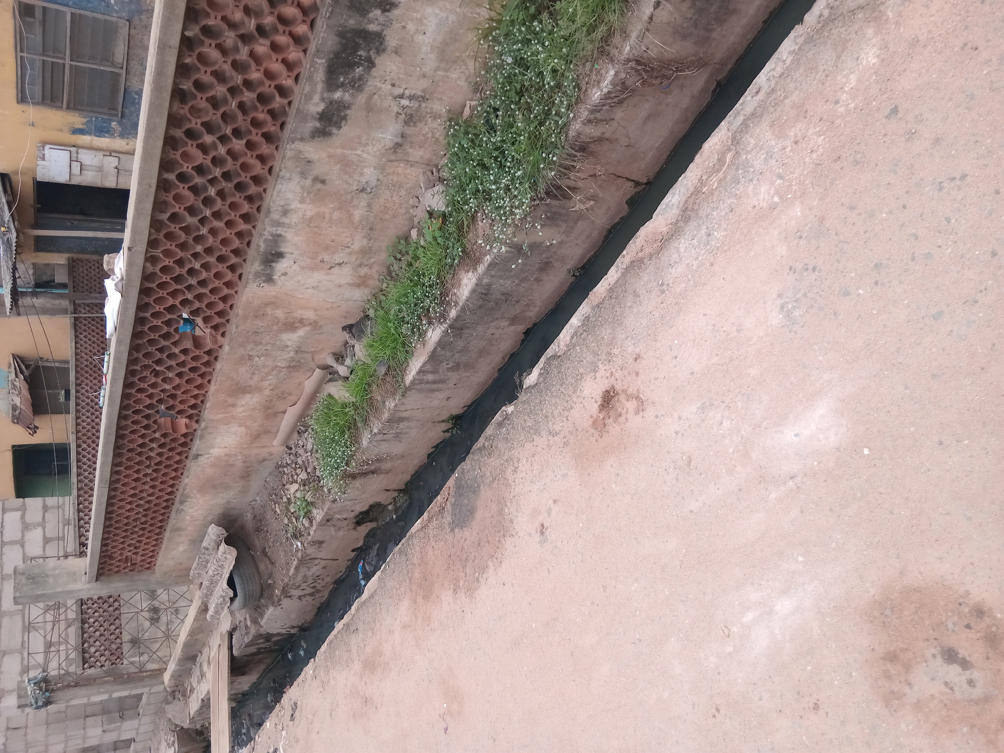 The image shows a narrow, open drainage ditch running alongside a worn, light-brown concrete road in a neighborhood in Shagamu, Nigeria. The ditch contains stagnant, dark water and some visible debris. Green vegetation grows along one edge of the ditch, closer to a building. The building is constructed with concrete blocks and features an open-lattice concrete design on its facade. Windows and laundry lines are visible on the building. The surrounding environment appears urban and somewhat neglected. A discarded tire is on the edge of the drainage.