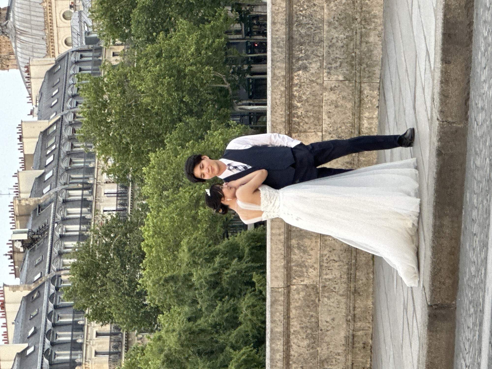 A newly married couple poses on a bridge in Paris, France. The bride is wearing a white wedding gown, and the groom is in a navy suit. The background features Parisian buildings and lush green trees. The couple are in an embrace.
