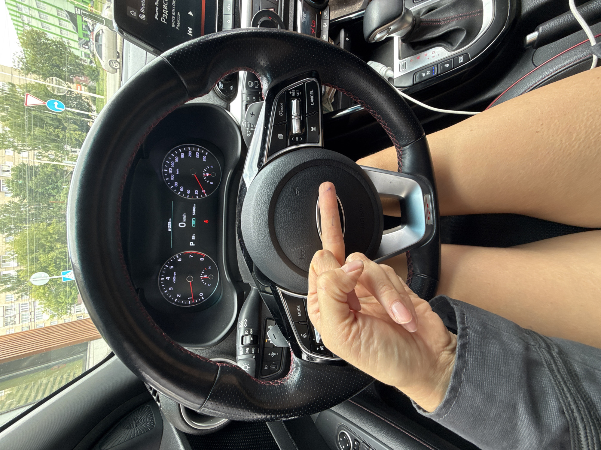The image shows a close-up view from inside a car, looking at the steering wheel and dashboard. A person's hand is positioned over the center of the steering wheel, with their index finger extended and appearing to touch the airbag cover. The dashboard displays various indicators, including speed, fuel level, and mileage which reads "57446 km". A phone screen is visible to the right of the steering wheel, displaying the text "iPhone АНС" and "Радио". The car appears to be parked or stopped, with traffic lights and buildings visible through the windshield, suggesting an urban setting. The overall scene is consistent with being in Izhevsk, Russia, given the Cyrillic text on the radio display. The lighting suggests it is daytime.