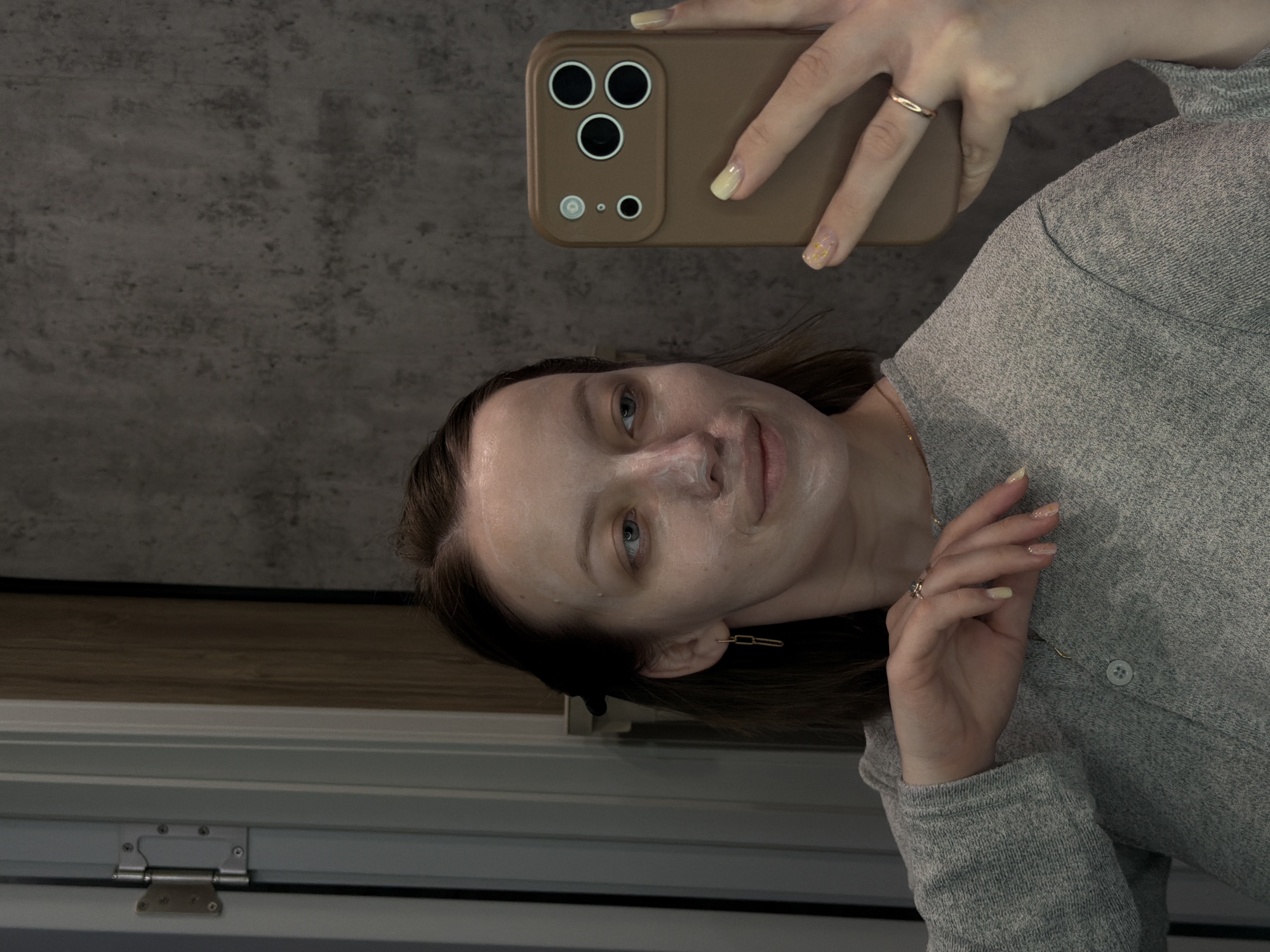 A young woman is captured taking a mirror selfie indoors, appearing to be in Reshetnikovo, Russia. She has a light-colored face mask applied to her face and looks towards the camera with a slight, calm smile. Her dark hair is pulled back from her face, revealing her blue-grey eyes.
She wears a light grey, long-sleeved top, a thin gold necklace, and a gold-toned ring on her left ring finger. A distinctive gold-toned paperclip-link earring hangs from her right ear. Her nails are painted light yellow, with some featuring small decorative elements. She holds a smartphone in a brown/taupe case with prominent camera cutouts, consistent with a recent iPhone model, to capture the image.
The background behind her head features a textured dark grey concrete wall. Below this, a strip of light brown wood, possibly a window sill, is visible above a white window or door frame with a silver hinge. The lighting suggests an indoor setting during daylight hours. No visible text is present in the image.