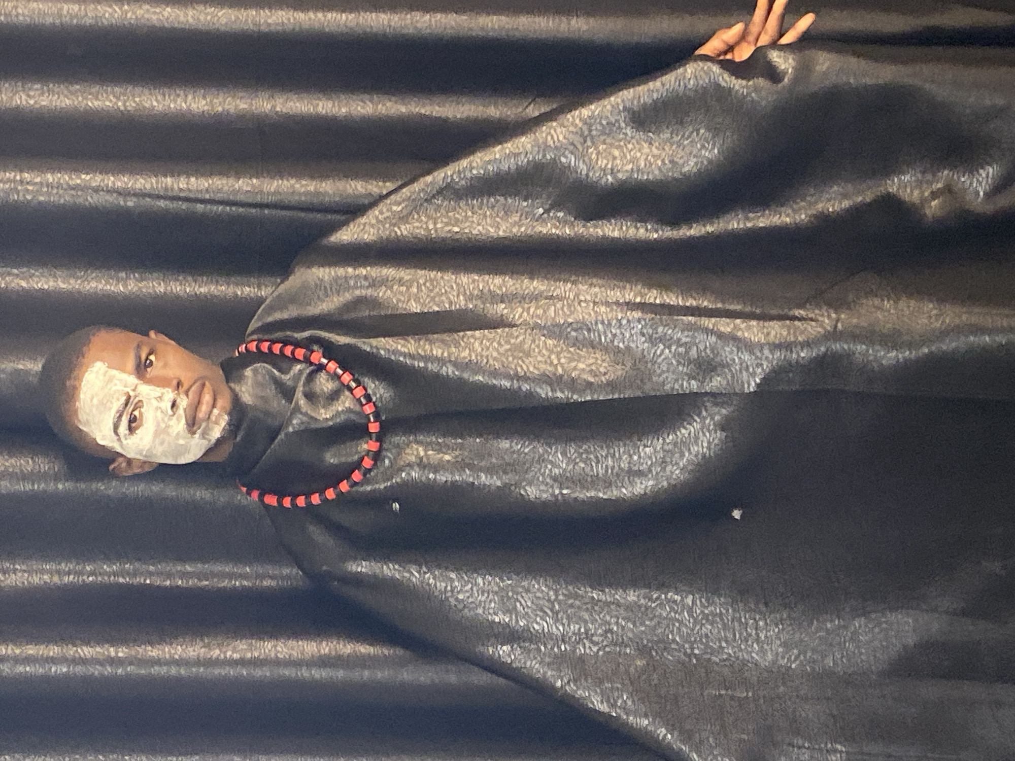 A young Black man stands before a dark, vertically pleated backdrop. His face is partially covered with white paint, with the right side of his face showing his natural skin tone. He wears a black cloak with a textured pattern and a red and black beaded necklace. His right hand is visible at the edge of the cloak, with fingers extended. The setting is likely indoors, given the backdrop, and the lighting suggests it is not natural daylight. There are no discernible landmarks or specific details that would indicate a location within Abakaliki, Nigeria.