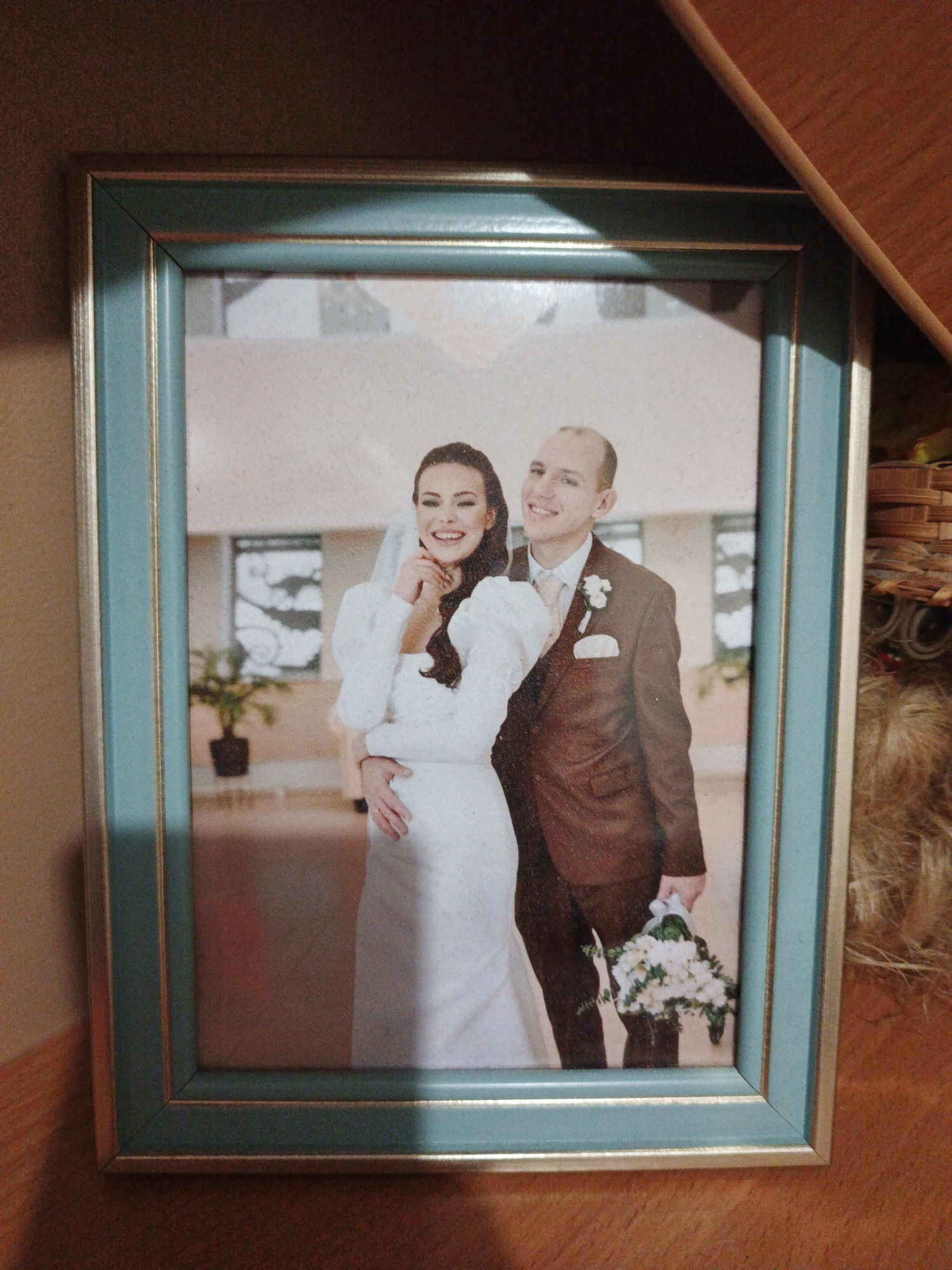 A framed photograph depicts a wedding portrait of a smiling Caucasian couple. The bride, on the left, wears a white wedding dress with puffed sleeves and a veil. The groom, on the right, is in a brown suit, white shirt, and tie, holding a bouquet of white flowers. They are positioned indoors, with a blurred background that includes a potted plant and architectural elements of a building. The photograph is enclosed in a teal and gold picture frame. The overall image quality suggests it is a physical photograph. The image does not contain any identifiable landmarks or features that would allow for the determination of its location in Kazan, Russia, or any other specific city.