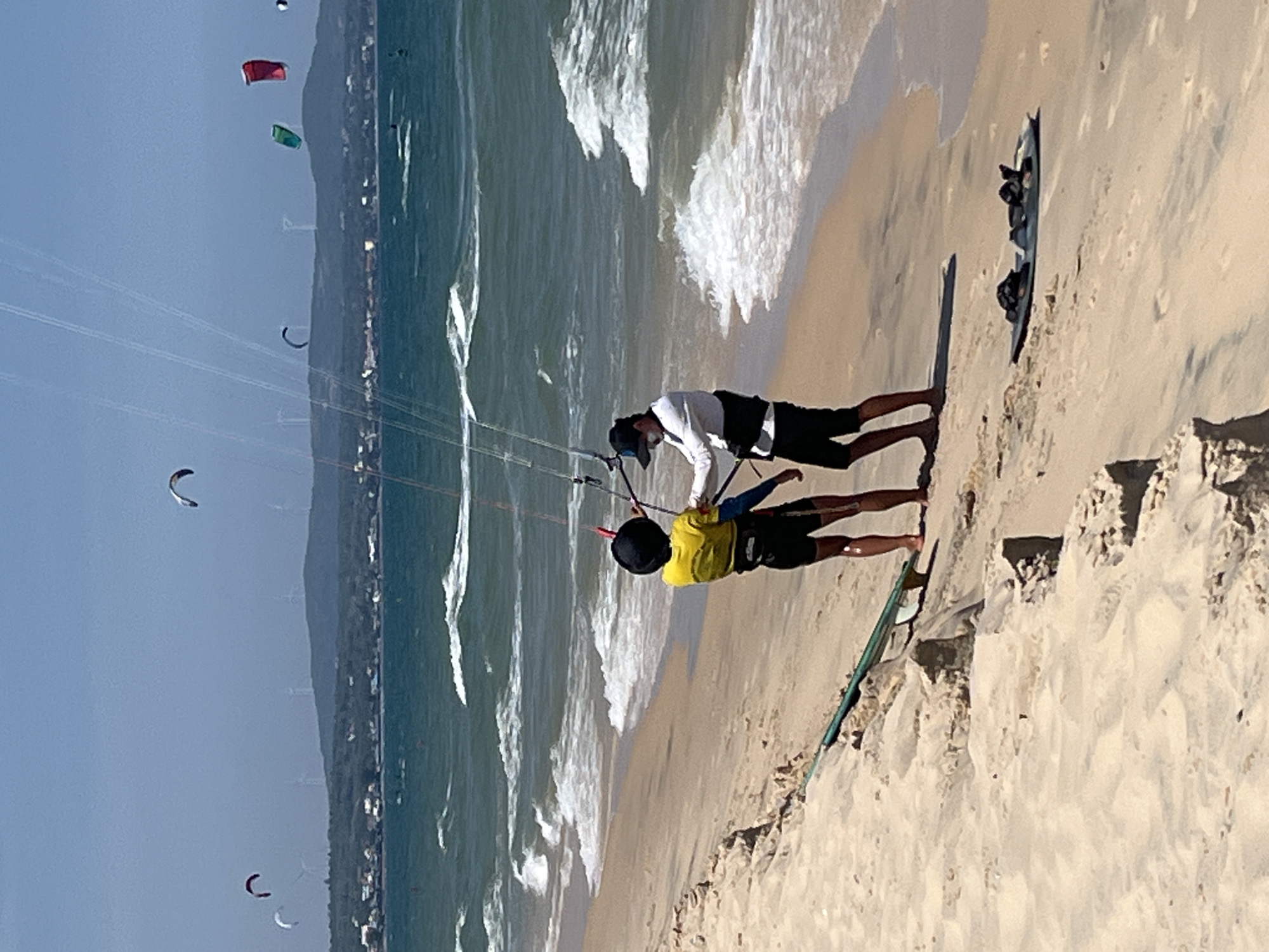 The image captures a sunny day at the beach in Phu Thuy Ward, Vietnam, with several kitesurfers in the ocean and two individuals on the sandy shore. One person, wearing a yellow t-shirt and black shorts, is holding onto the kite lines. The second person, dressed in a white long-sleeved shirt and black shorts, is assisting them. A kiteboard lies on the sand near them, along with another board further down the beach. In the background, the turquoise ocean is dotted with several colorful kites, indicating active watersports. The coastline features a sandy beach with some vegetation and a distant view of a town with wind turbines on a hill under a clear blue sky. The overall scene depicts a vibrant and active kitesurfing location.
