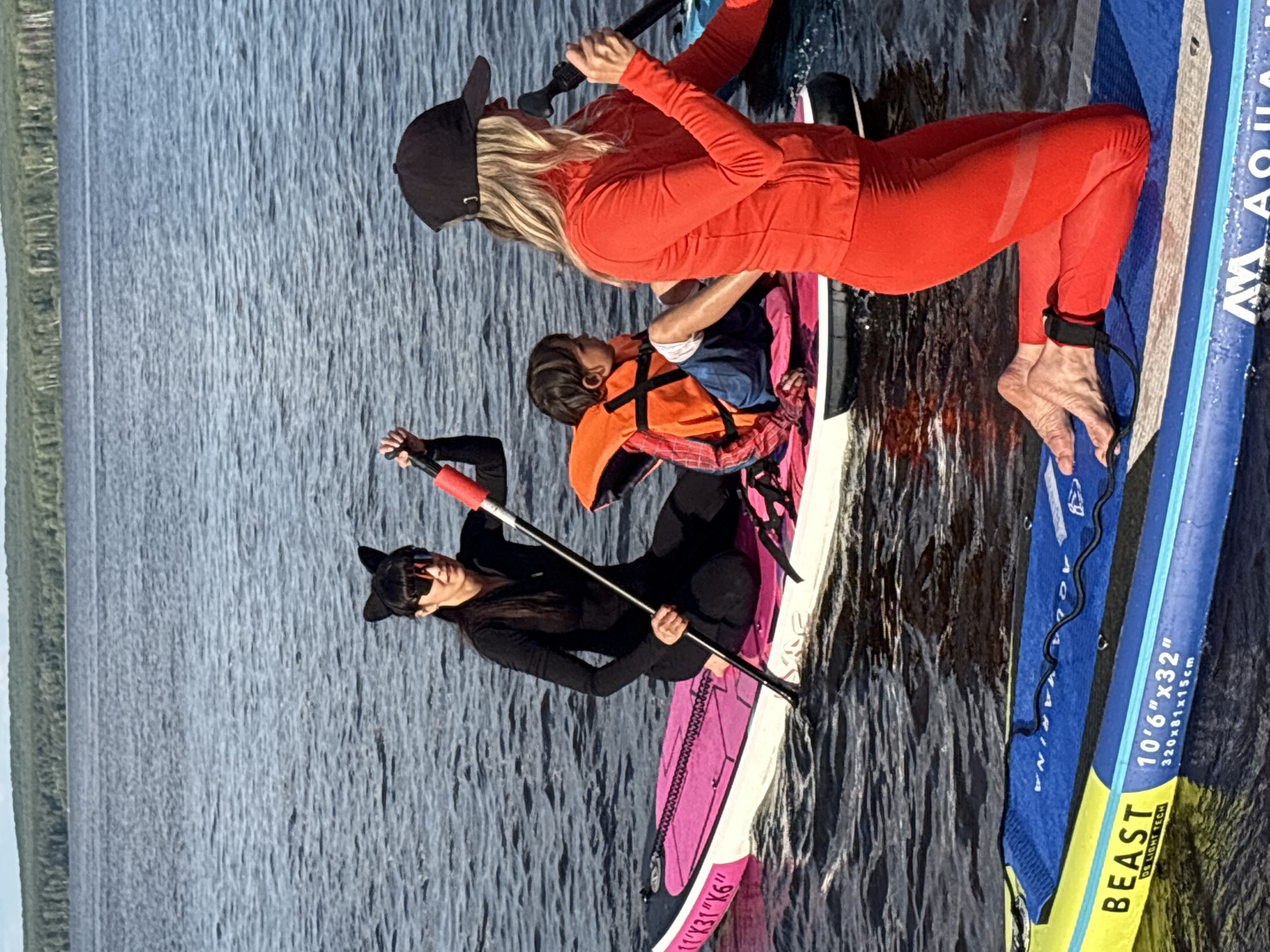 In this outdoor, eye-level shot, three people are paddleboarding on a body of water. The person in the foreground, dressed in a black long-sleeved shirt and black leggings, is kneeling on a pink and white paddleboard, holding a paddle. They are wearing black cat ears and sunglasses. Behind them, another person is seated on the same paddleboard, wearing an orange life vest over a blue shirt. This person is also wearing a black baseball cap. Further back, a third person is standing on a blue paddleboard, wearing a bright orange long-sleeved shirt and red leggings. They are also wearing a black baseball cap and holding a paddle. The water is a deep blue with visible ripples, suggesting a calm day. The background shows a blurred line of trees along the shore. The lighting indicates it is daytime, likely mid-morning or afternoon. The paddleboards are branded with text including "BEAST," "A MARINA," and dimensions.