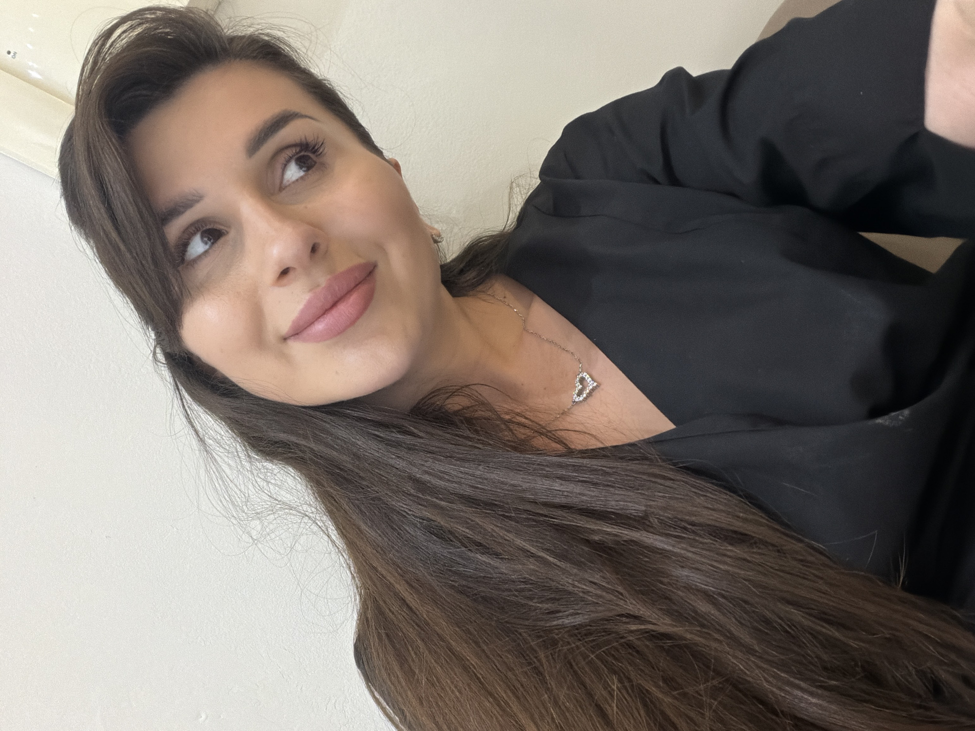 A selfie-style portrait features a woman with long, dark brown hair parted on the side, cascading over her shoulder and chest. She is looking upwards and to the right with a soft, slightly smiling expression, her lips painted with light pink lipstick. Her skin appears smooth, and she has visible eyelashes and shaped eyebrows. She is wearing a black top or dress, and a delicate silver necklace with a heart-shaped pendant adorned with small gems is visible on her décolletage.
The background is a plain, light-colored (likely white or off-white) wall. In the upper left corner, a white fixture, possibly a vent or light switch cover, is partially visible, with faint text reading "ON" and "OFF". The lighting suggests an indoor setting. The woman's pose and direct gaze away from the camera suggest a moment of reflection or a posed shot. The overall impression is one of calm.
The setting for this image is Budva, Montenegro.