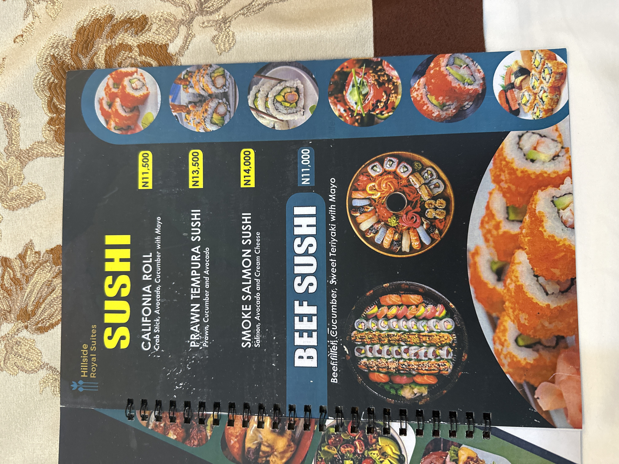 The image is a close-up, overhead shot of a menu from Hillside Royal Suites. The menu features a variety of sushi dishes with prices listed in Nigerian Naira (N). The dishes include "CALIFORNIA ROLL" for N11,500, "PRAWN TEMPURA SUSHI" for N13,500, "SMOKE SALMON SUSHI" for N14,000, and "BEEF SUSHI" for N11,000. Each dish is accompanied by a small circular photograph showcasing the sushi. The menu is bound with a black spiral binding. The background of the image is a textured, light brown fabric. There are no people visible in the image. The location of the Muhammadu Dikko Stadium in Nigeria is not mentioned or depicted in this image.