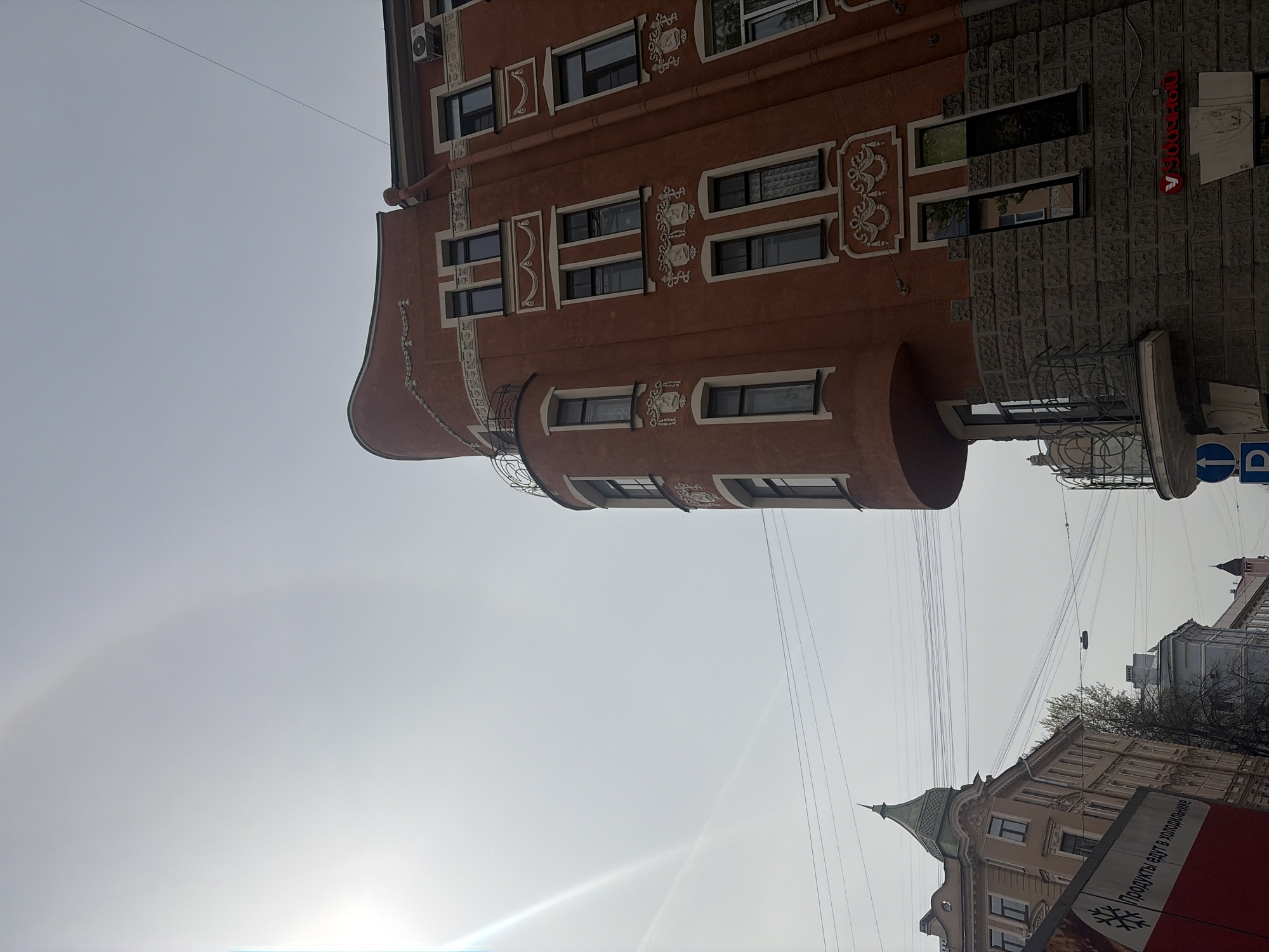 The image shows an exterior view of a building in Saint Petersburg, Russia. The architectural style features decorative window frames, a curved corner facade, and a terracotta color. A sign in Cyrillic is visible on the building. Overhead wires crisscross the sky. Another building with a spire is partially visible in the background. The sky is bright, suggesting daytime.