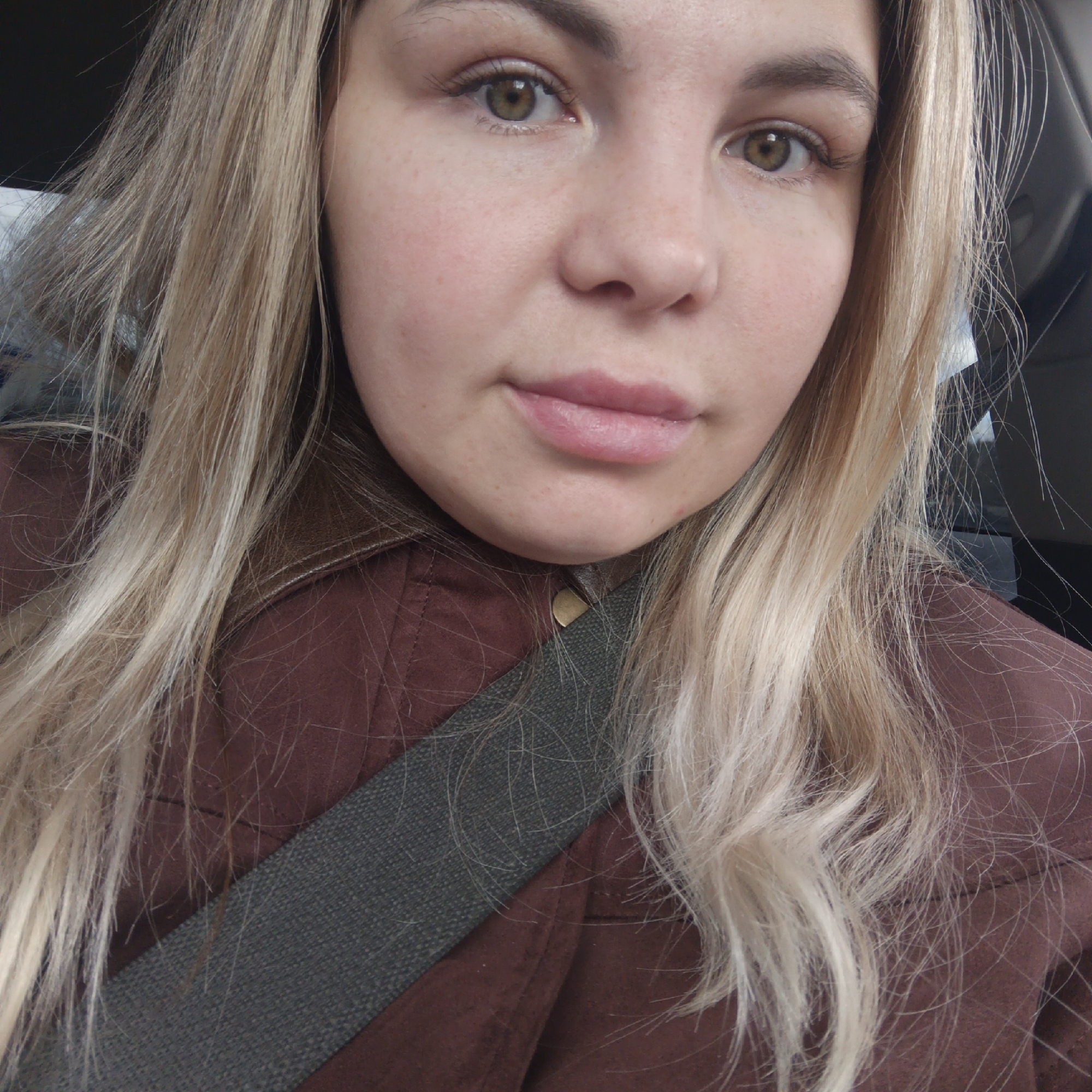 A close-up portrait shows a young woman with long, light blonde hair looking directly at the camera. Her hair, which appears to be styled straight to slightly wavy, frames her face and extends down over her shoulders. She has fair skin, with a few visible freckles on her nose and cheeks. Her eyes are a striking hazel or greenish-brown color, and her expression is neutral with a hint of a soft smile. She appears to be wearing minimal makeup, with possibly some light mascara and lip balm or gloss on her lips.
She is dressed in a brown jacket or coat, which has a structured collar and a visible golden button near her neck. A grey, textured seatbelt diagonally crosses her torso, suggesting she is inside a vehicle, likely a car. The background is blurred and dark, consistent with a vehicle interior. The lighting on her face suggests it is daytime. There is no visible text in the image.