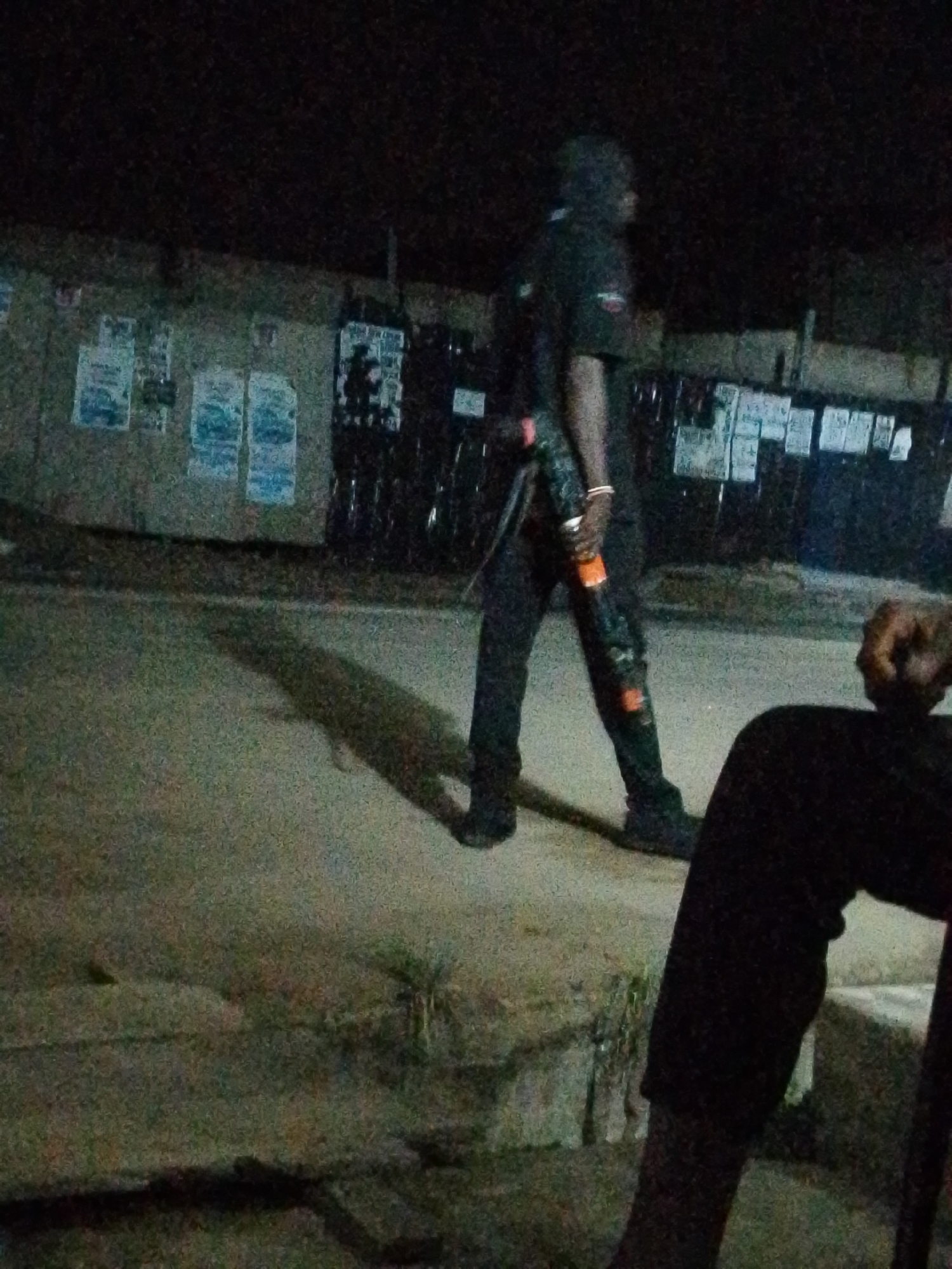 The image was taken at night in Ifite-Awka, Nigeria. A man dressed in dark clothing is walking across the street, carrying a can of drink in his hand. The location appears to be an outdoor space, with buildings or structures behind him. There are posters attached to the structure. In the foreground, a leg is sitting, perhaps sitting on a chair.