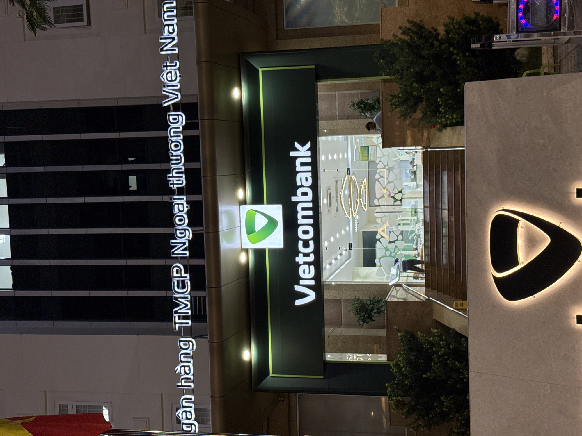 The image shows the exterior of a Vietcombank branch in Nha Trang, Vietnam, at night. The bank's signage is prominently displayed, with "Vietcombank" in large white letters on a dark green background. Above this, a sign reads "Ngân hàng TMCP Ngoại thương Việt Nam," which translates to "Joint Stock Commercial Bank for Foreign Trade of Vietnam." A green triangular logo is also visible.
The entrance to the bank features a set of stairs leading up to the doors. Inside, the bank appears to be well-lit with modern lighting fixtures. A few people are visible: one individual, possibly a security guard or employee, is standing near the entrance. Another person is seated inside the bank, looking out.
The building itself is modern, with multiple stories and dark vertical window frames. A Vietnamese flag, predominantly red with a yellow star, is visible on the left side of the image, attached to a flagpole. The surrounding area is dark, indicating nighttime. There are some trees and bushes visible in front of the bank.
