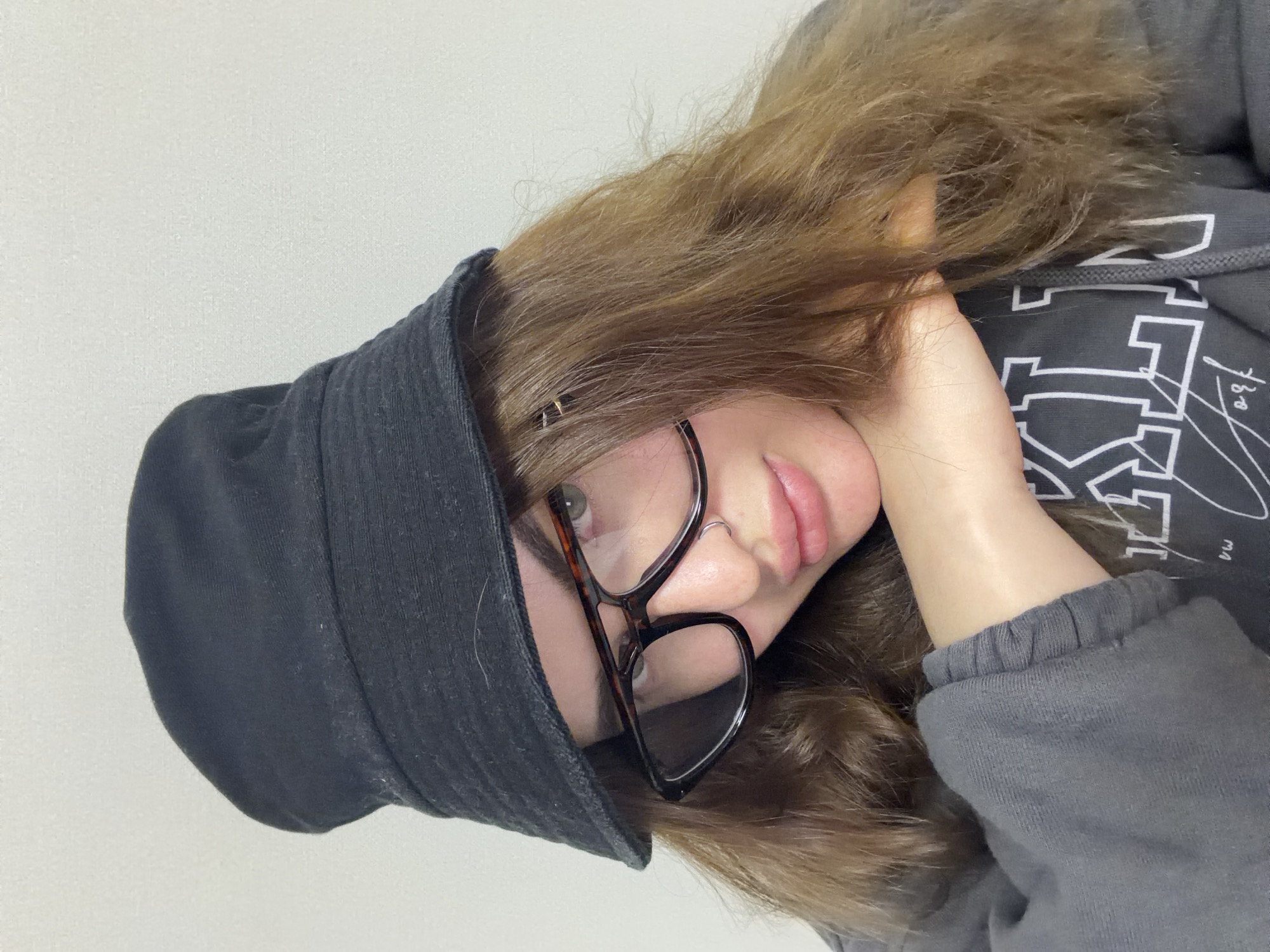 A close-up portrait features a young person, likely female, with long, wavy light brown hair. The subject is wearing dark-framed eyeglasses and a dark, possibly black, bucket hat. Her head is tilted, and she looks directly towards the camera with a neutral, calm expression, her lips slightly parted. A small ring piercing is visible on her right nostril.
She is dressed in a dark grey hooded sweatshirt (hoodie), the ribbed cuff of which is visible on her left arm as it rests near her face. The front of the hoodie displays a partially visible white graphic design, featuring stylized block letters that appear to spell out "PINK" or a similar brand, along with faint lines and smaller text such as "180" and "m3". The background is a plain, light-colored (white or off-white) wall, indicating an indoor setting. This image is from Krasnodar, Russia.