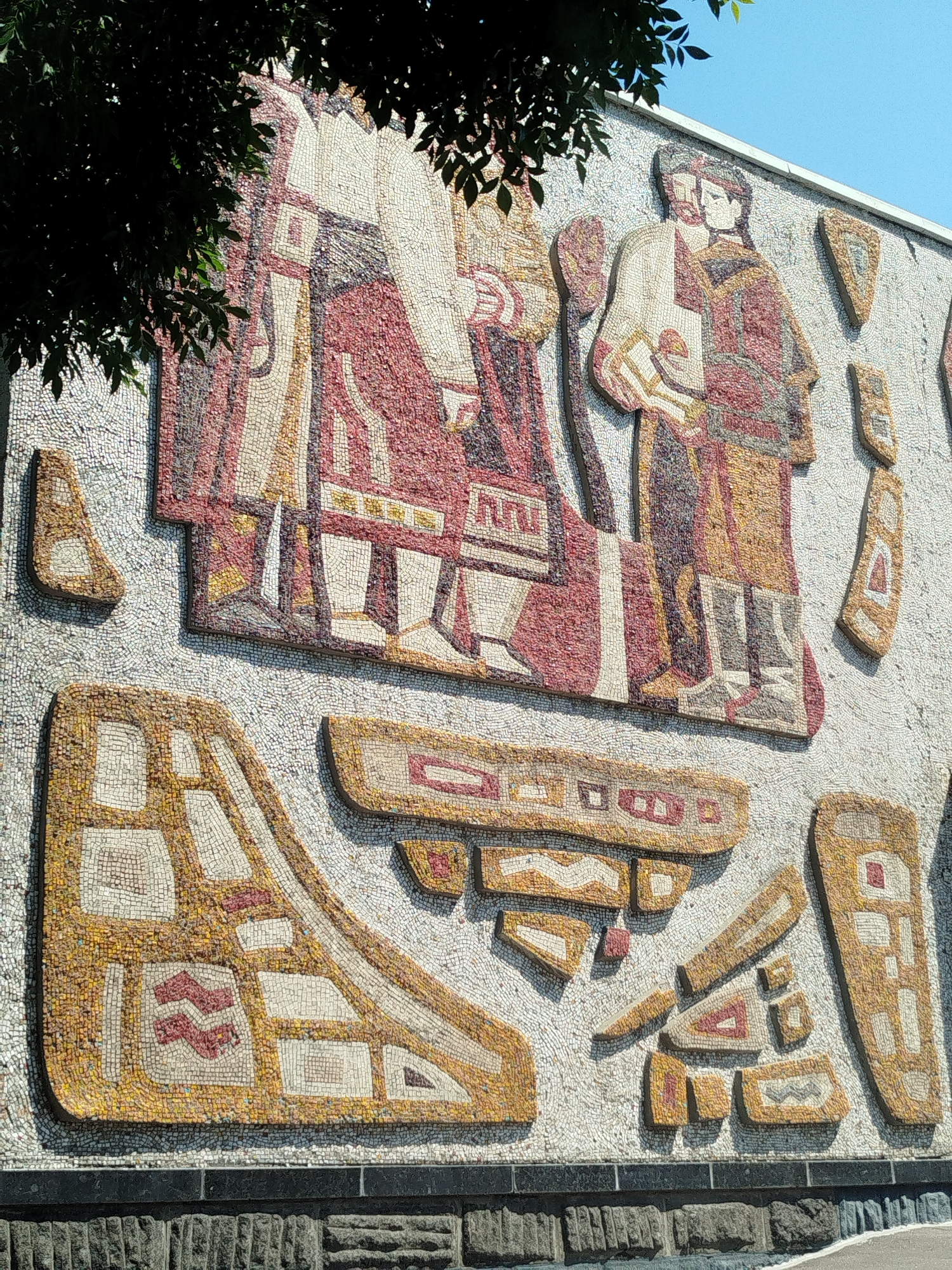This image displays a large, intricate mosaic mural on the exterior wall of a building in Kremenchuk, Ukraine. The mosaic depicts figures in traditional clothing, characterized by rich red, beige, and yellow tones, set against a white background composed of countless small tiles. The mural features two central figures standing side-by-side, with one appearing to hold an object. Below these figures, abstract geometric shapes and patterns are arranged in a mosaic style, also in similar color palettes. The artwork is mounted on a building with a base constructed from dark stone blocks. A tree with dark green foliage partially obscures the upper left portion of the mosaic. The overall impression is of a historically or culturally significant public art installation.