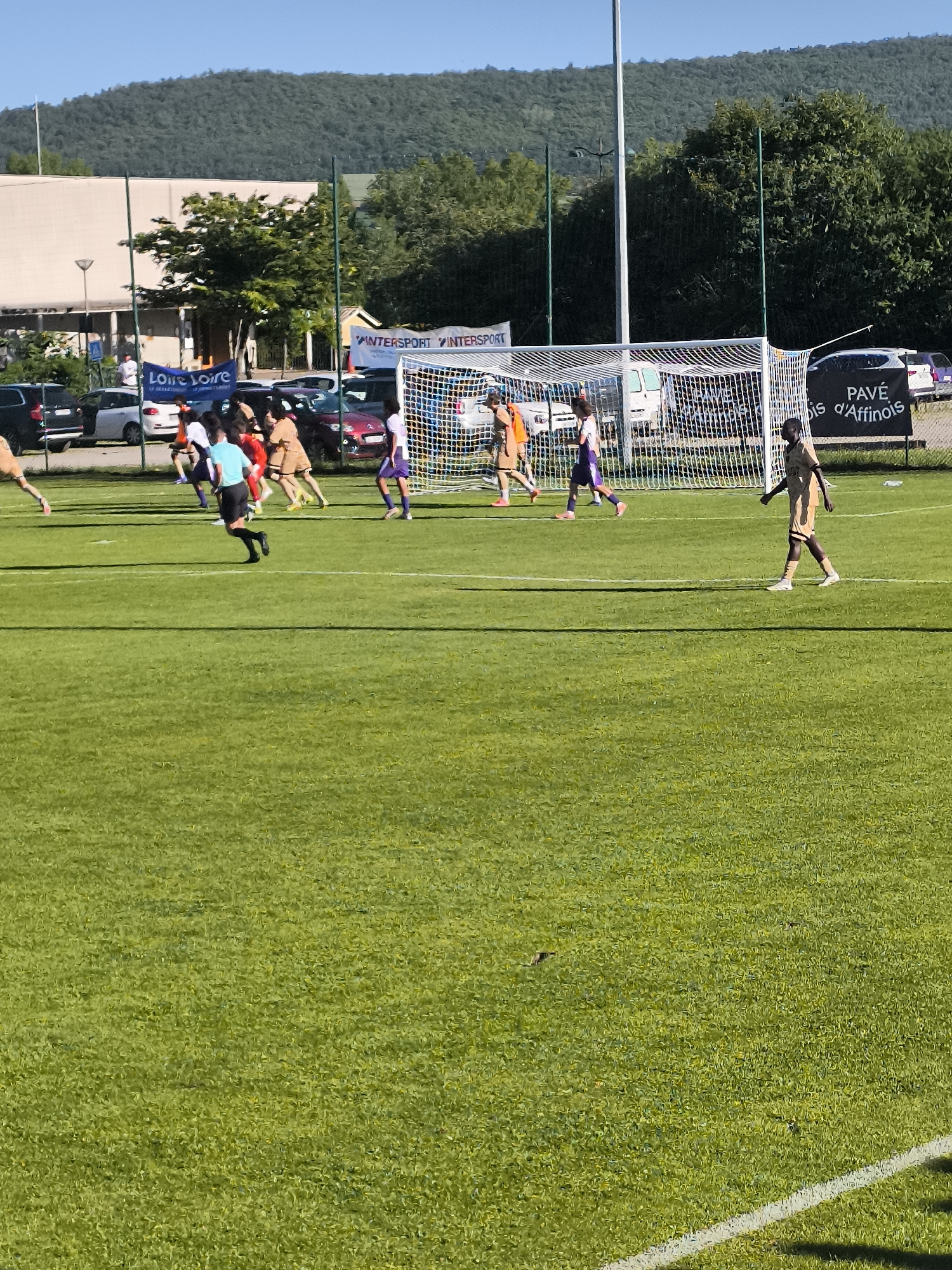 The image shows a soccer game in progress on a green field. Players in various uniforms (orange, purple, white, and gold/brown) are visible near the goal. A referee in black shorts and a blue shirt is also present. In the background, there are parked cars, banners with the words "INTERSPORT" and "LOIRE LOIRE" on them, and a banner that says “PAVE is d’Affinois”. A building is visible in the background, along with trees and a mountain range. Based on the banner, the image is located in Maclas, France.