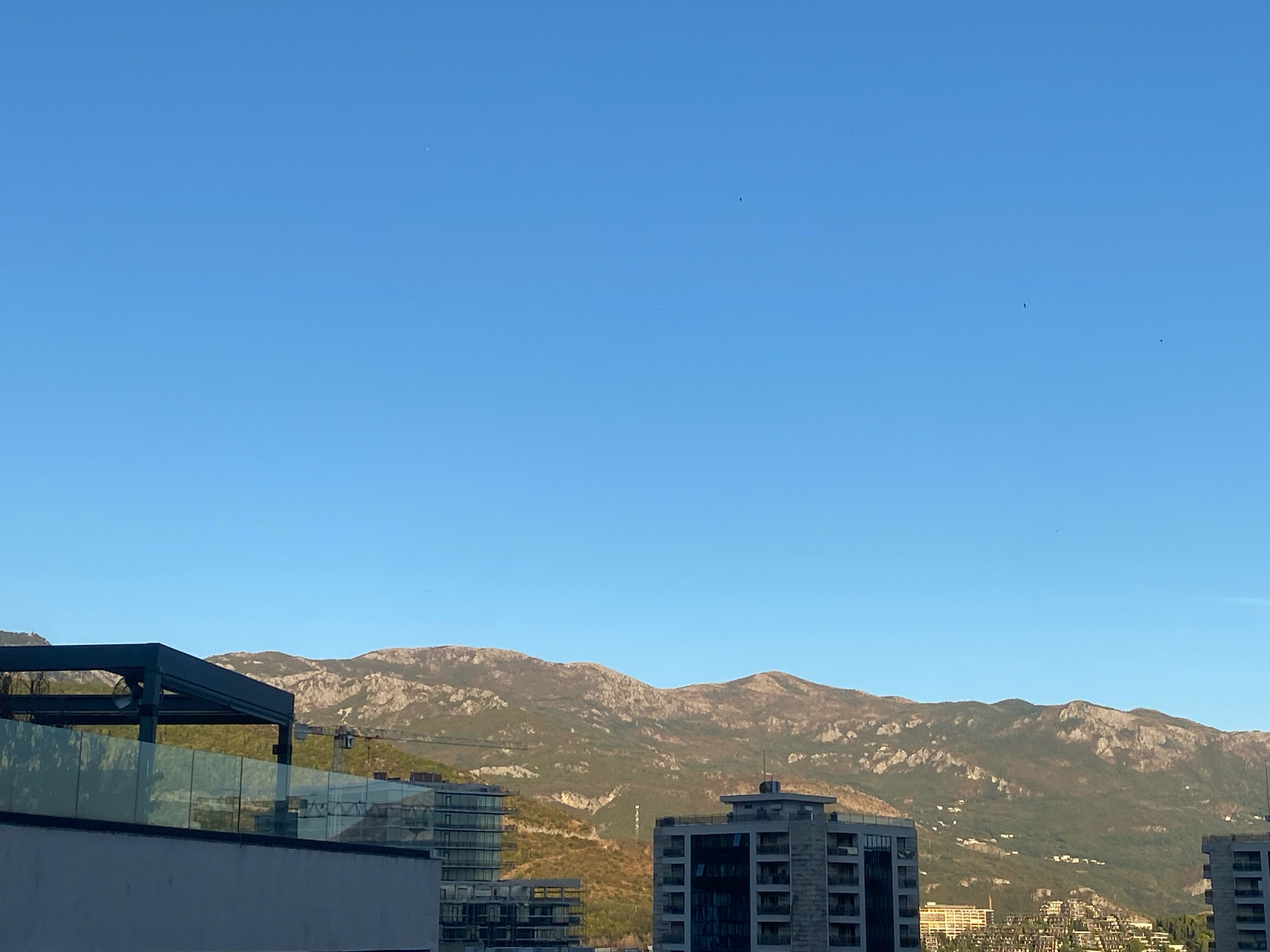 The image captures a cityscape with a backdrop of mountains under a clear blue sky. In the foreground, a modern building with a glass balcony railing is partially visible. Behind it, several multi-story buildings, some appearing to be residential or hotel complexes with glass facades, are situated. A construction crane is also visible amongst the buildings. The mid-ground is dominated by rolling hills and rugged mountains, some of which are covered in sparse vegetation and rocks. Small clusters of buildings are scattered on the lower slopes of the mountains. The overall scene suggests an urban environment in a hilly coastal region, likely Budva, Montenegro, given the characteristic architecture and mountainous terrain. The lighting indicates it is daytime, possibly late afternoon or early morning, with the sun casting soft shadows. There are no people or animals clearly depicted in the image.