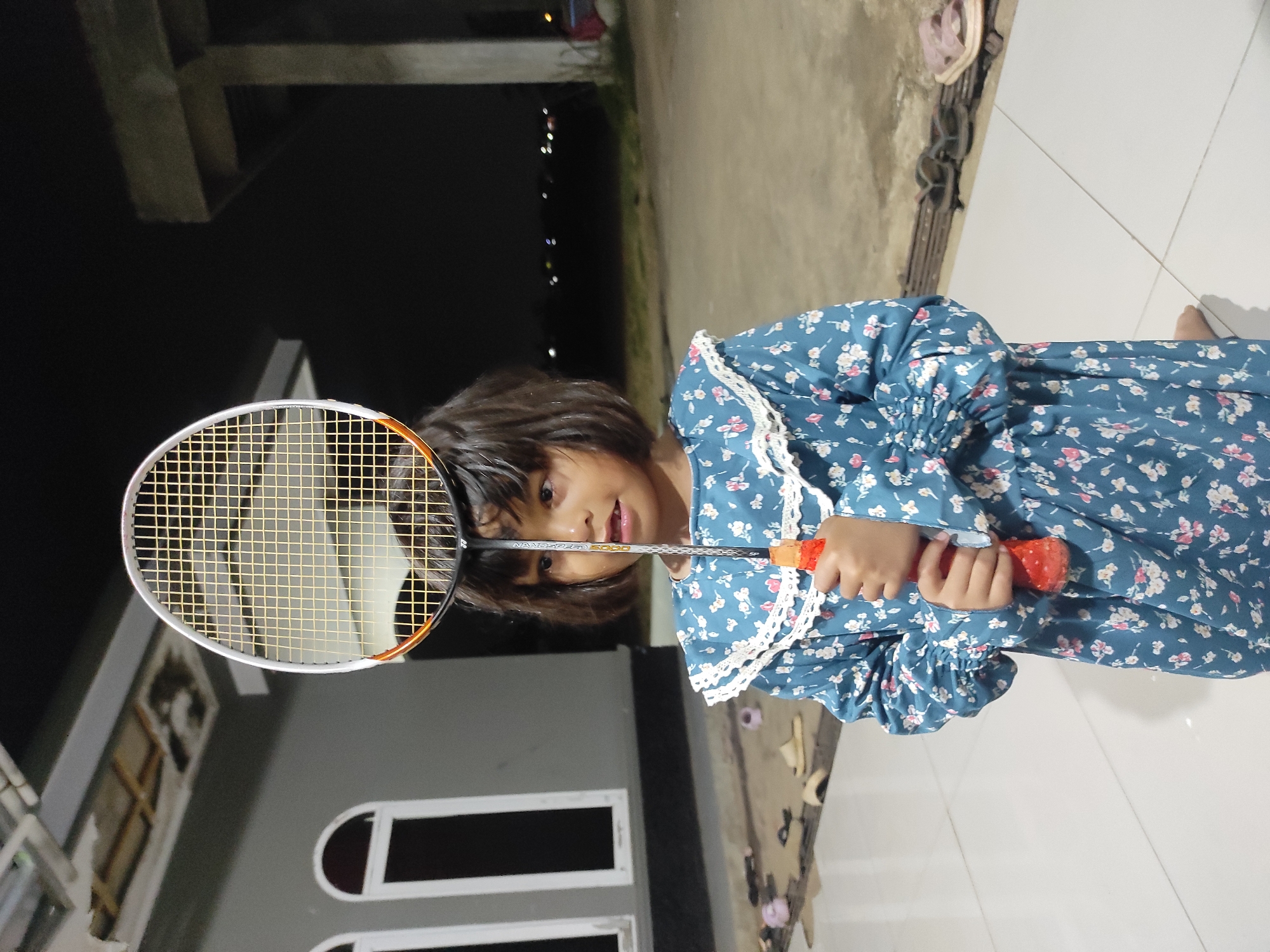 A young girl stands outdoors at dusk, holding a badminton racket vertically in front of her face. She is wearing a blue floral dress with a white lace collar and cuffs. The girl has dark hair and is looking slightly to her left with her mouth open. The background shows a grey building with arched windows, and beyond that, a dark, possibly rural, landscape with scattered lights. The ground is tiled in the foreground, with a concrete patio area visible further back. Several pairs of sandals are scattered on the ground in the background. The overall lighting suggests it is nighttime or late evening.