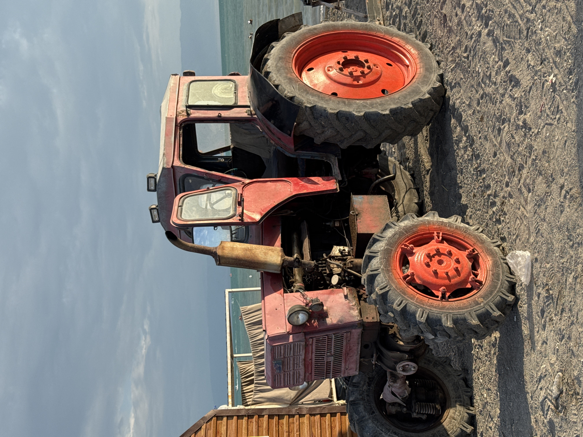 A weathered red tractor sits on a sandy beach with the ocean in the background. The tractor is a Soviet-era model, likely from the 1970s or 1980s, and shows signs of age and use. Its paint is faded, and there are visible rust spots and dirt. The tractor has large, rugged tires, a tall exhaust pipe, and a cab with open doors and windows. To the left of the tractor is a portion of a wooden structure, possibly a small building or shed. In the distance, the ocean stretches out to the horizon under a cloudy sky. The overall scene suggests a rural or coastal location. The provided location of Zharbulak, Kazakhstan, places this scene in a specific geographic context, implying a dry or continental climate, potentially near a lake or a smaller sea if not a large ocean. The presence of the tractor on a beach could indicate its use for maintenance or transport in a waterside area, or it could simply be parked there. There are no people visible in the image.
