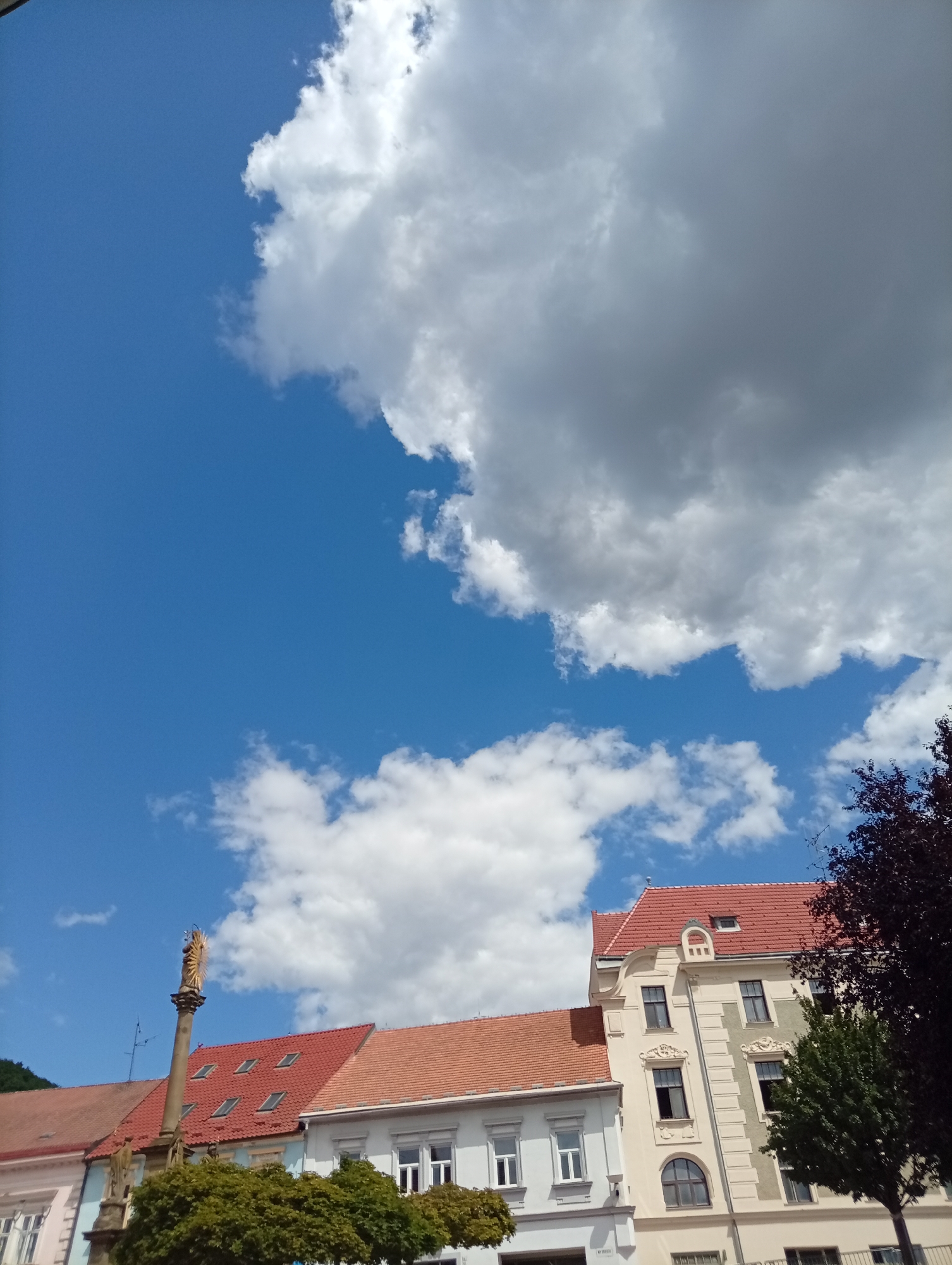 The image shows a clear blue sky with large, fluffy white and gray clouds on a bright, sunny day. In the lower half of the frame, several buildings with red tiled roofs are visible, along with a tall column topped with a golden statue. A large, leafy green tree is in the foreground. The buildings appear to be located in a town square, as suggested by the presence of the statue and the architectural style. The scene is likely in Tišnov, Czechia, given the reference to that location. No people are visible in the image.