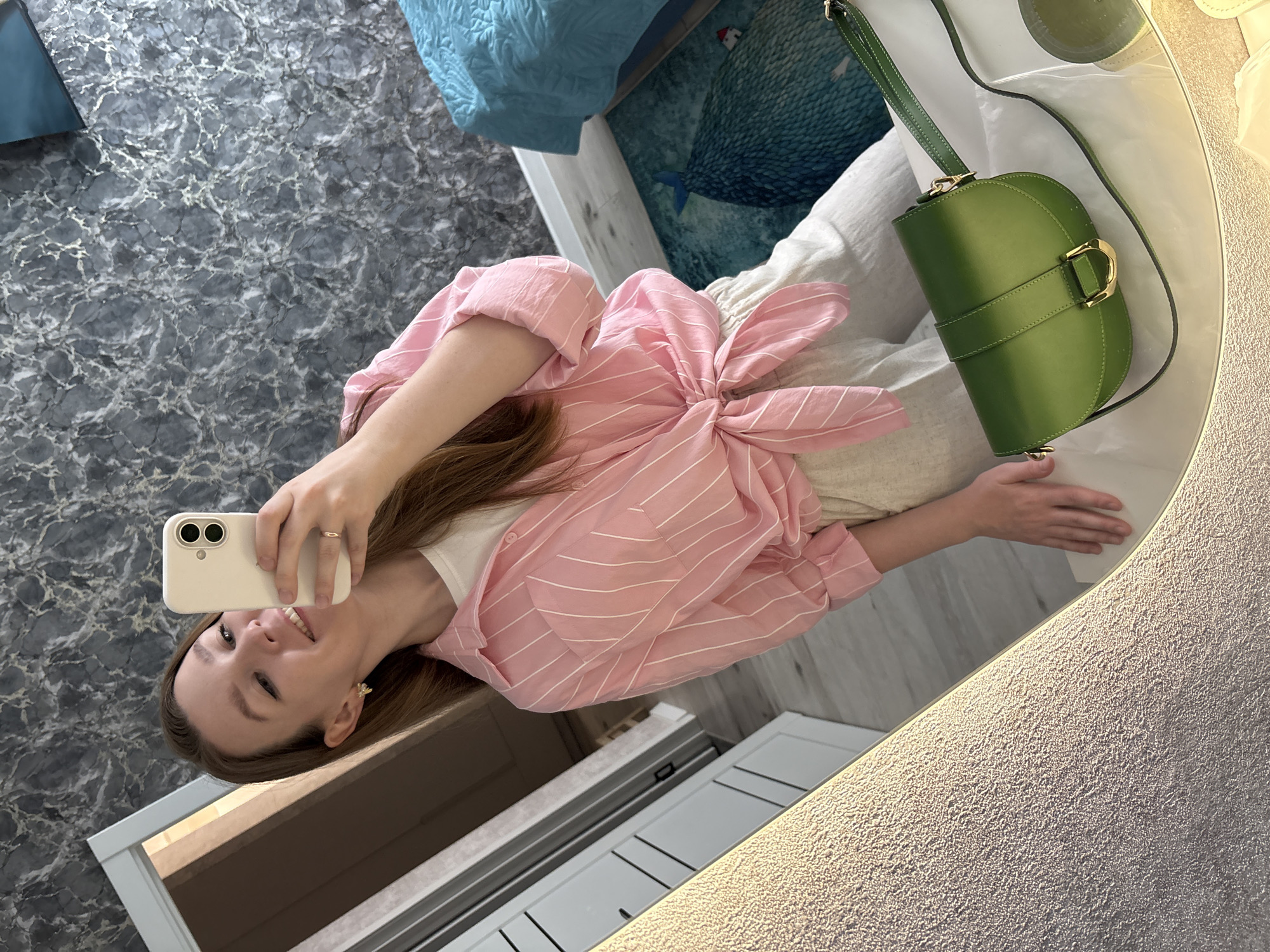 A woman is taking a selfie in a mirror. She is wearing a pink striped shirt tied at the waist and beige pants. The woman is smiling and holding a white smartphone in her left hand. Her right hand is resting on the edge of the mirror. A green handbag is on the surface next to her. A blue and green decorative bedspread is visible in the background. The mirror is framed by a textured gray material with a light strip. The scene appears to be indoors, possibly in Izhevsk, Russia, but there is insufficient evidence to confirm the location.