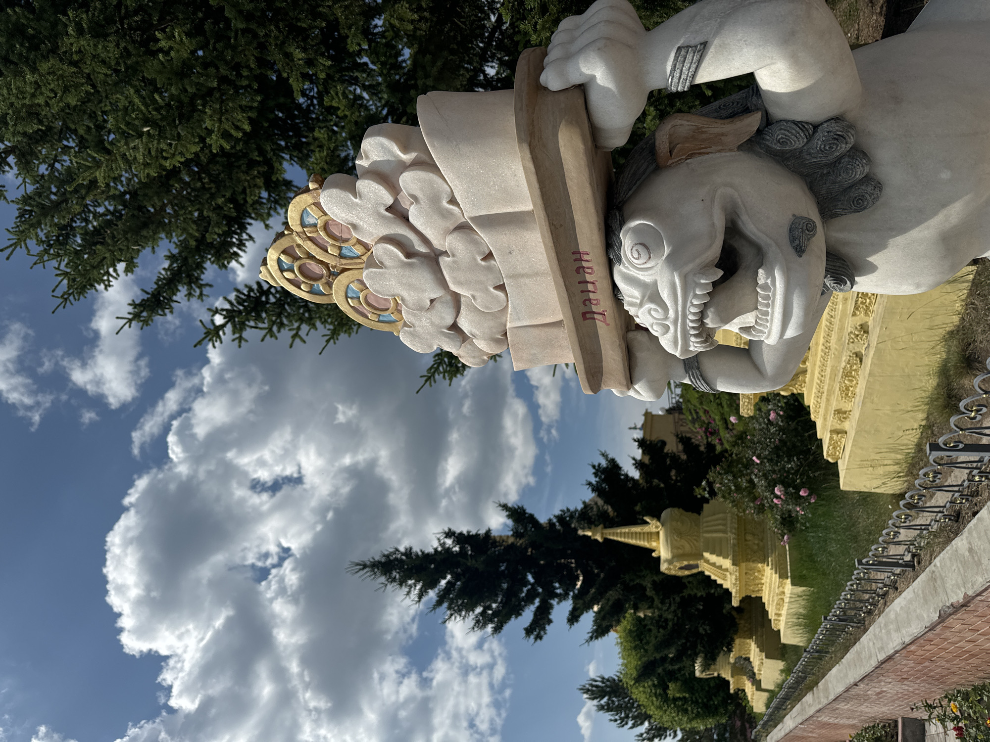 The image depicts a sculpture of a mythical creature with its mouth open, holding a large, ornate, cloud-like object on its head. The sculpture is made of white stone and has intricate carvings. In the background, there are several golden stupas, trees, and a blue sky with white clouds. The scene appears to be in a temple or spiritual site. This photograph was taken at the Ivolginsky Datsan, the center of Buddhism in Russia, located near Ulan-Ude.