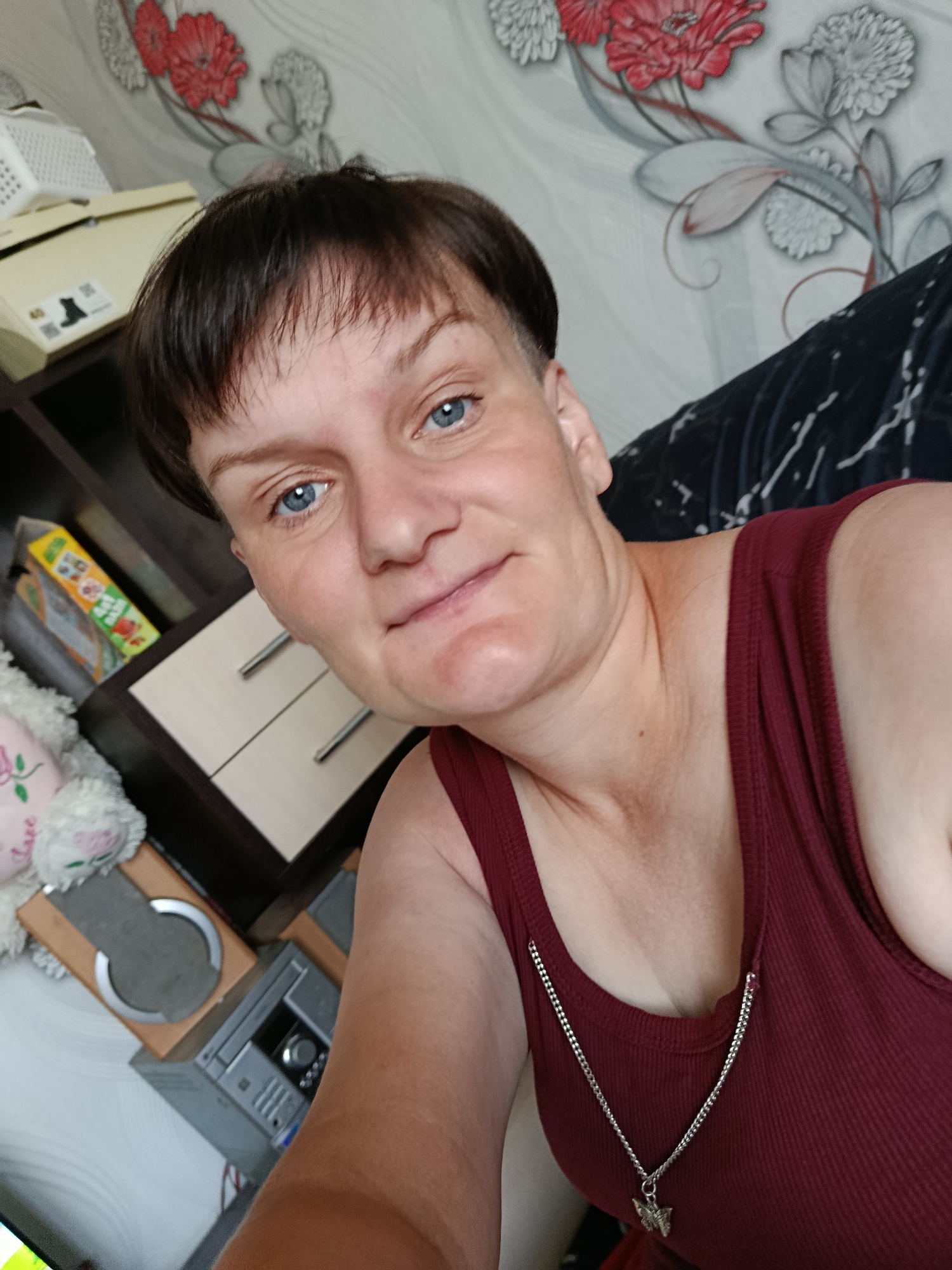 The image shows a self-portrait of a woman with short, dark brown hair and blue eyes. She is wearing a maroon tank top with a silver chain necklace that has a butterfly pendant. The background appears to be an indoor setting, possibly a home. There are shelves in the background with various objects, including a white storage container, a box with graphics, a stuffed animal, and what looks like a stereo system. The wallpaper in the background has a floral pattern with red and gray flowers. The woman's pose suggests she is looking directly at the camera. There is no indication of the location being in the city of Fokino, Russia.