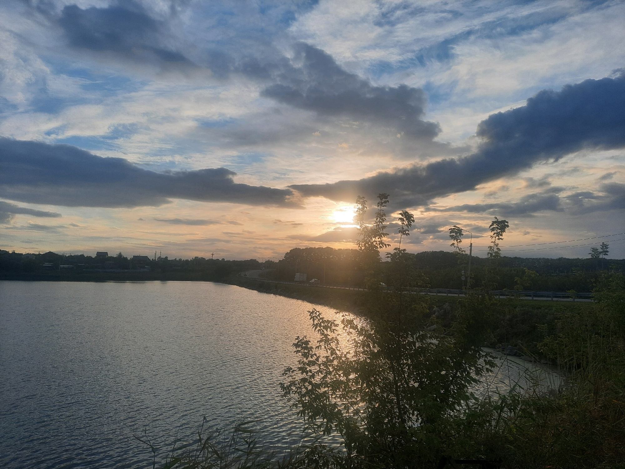 The image shows a serene sunset over a body of water, likely a lake or reservoir, near the city of Nauchny Gorodok, Russia. The sky is filled with a mix of dark and wispy clouds, with the setting sun casting a warm, golden glow through a break in the clouds. The water's surface reflects the sky, creating a shimmering effect. Along the far bank of the water, a treeline is visible, with houses and structures of the city of Nauchny Gorodok nestled behind the trees. A road runs along the edge of the water, with a few vehicles visible, suggesting a suburban or semi-rural setting. The foreground is dominated by vegetation, including tall grasses and leafy bushes, some of which are silhouetted against the bright sky. The overall impression is one of tranquility and natural beauty, with hints of human settlement in the distance.