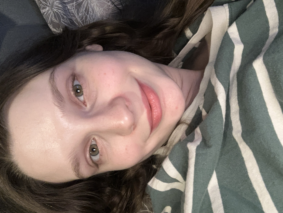 A woman lies with her head tilted, nestled amidst a gray patterned pillow and a green and white striped fabric. Her face is prominent, displaying fair skin, green eyes, and slightly parted lips. Dark brown hair frames her face and adds to the casual, relaxed ambiance of the image. She gazes directly at the viewer with a slight smile.