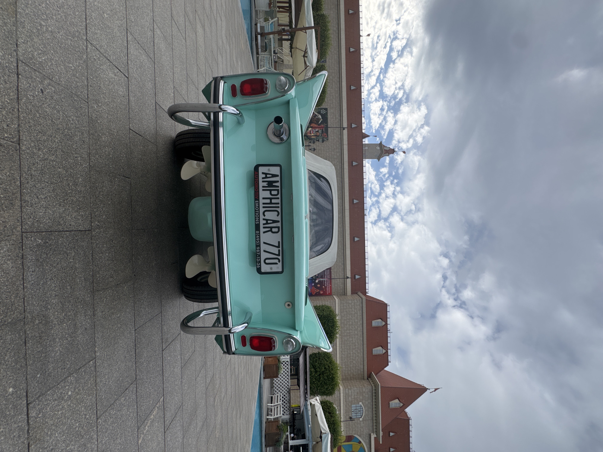 A rear view of a mint green Amphicar 770 is captured on a cloudy day in Moscow, Russia. The amphibious vehicle is parked on a paved walkway, with its two propellers visible at the back. The license plate reads "AMPHICAR 770".
In the background, a building with red-tiled roofs and stone walls stands under a sky filled with dramatic clouds. The architecture suggests a European influence, possibly a tourist attraction or a themed establishment. There are some outdoor seating areas with umbrellas and chairs visible, indicating a public space. The overall impression is that of a leisure or exhibition setting. The light suggests it's daytime, but the heavy cloud cover might make it appear overcast. There are no people visible in the frame.