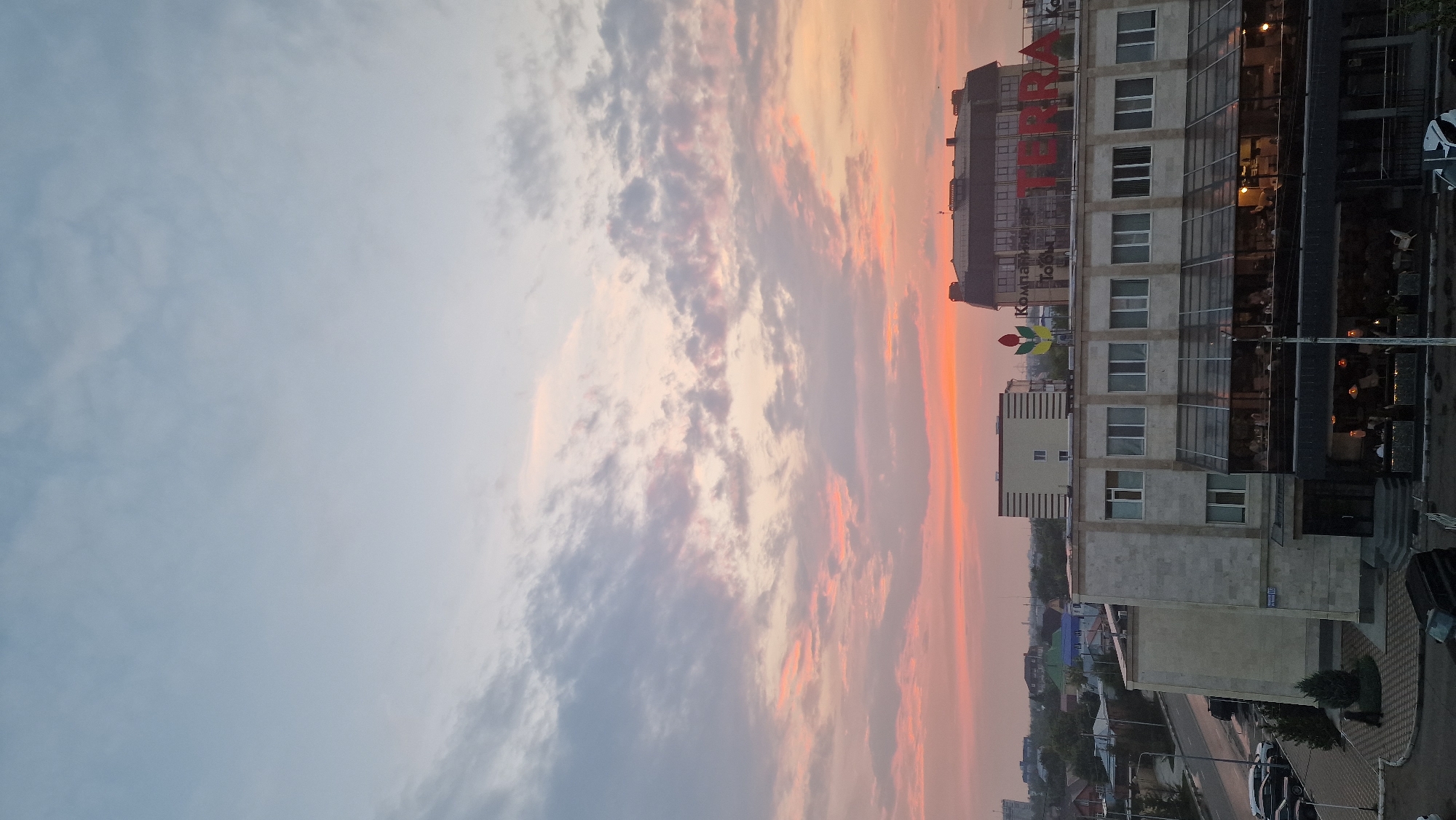 This media file depicts a cityscape during a sunset. The sky is filled with dramatic clouds painted in hues of orange, pink, and purple, indicating the golden hour. In the foreground, a modern multi-story building stands prominently. The building has a glass facade on its lower floors, revealing the warm interior lights and the silhouettes of people inside, suggesting a restaurant or commercial space. The upper floors are constructed with concrete and feature numerous windows. A large, red sign on the top of the building reads "TERRA". Below it, another sign with a logo featuring a flower shape and some text in Cyrillic letters is visible. The street in front of the building is lined with parked cars and some greenery. The overall scene evokes a sense of calm and beauty as day transitions into night.