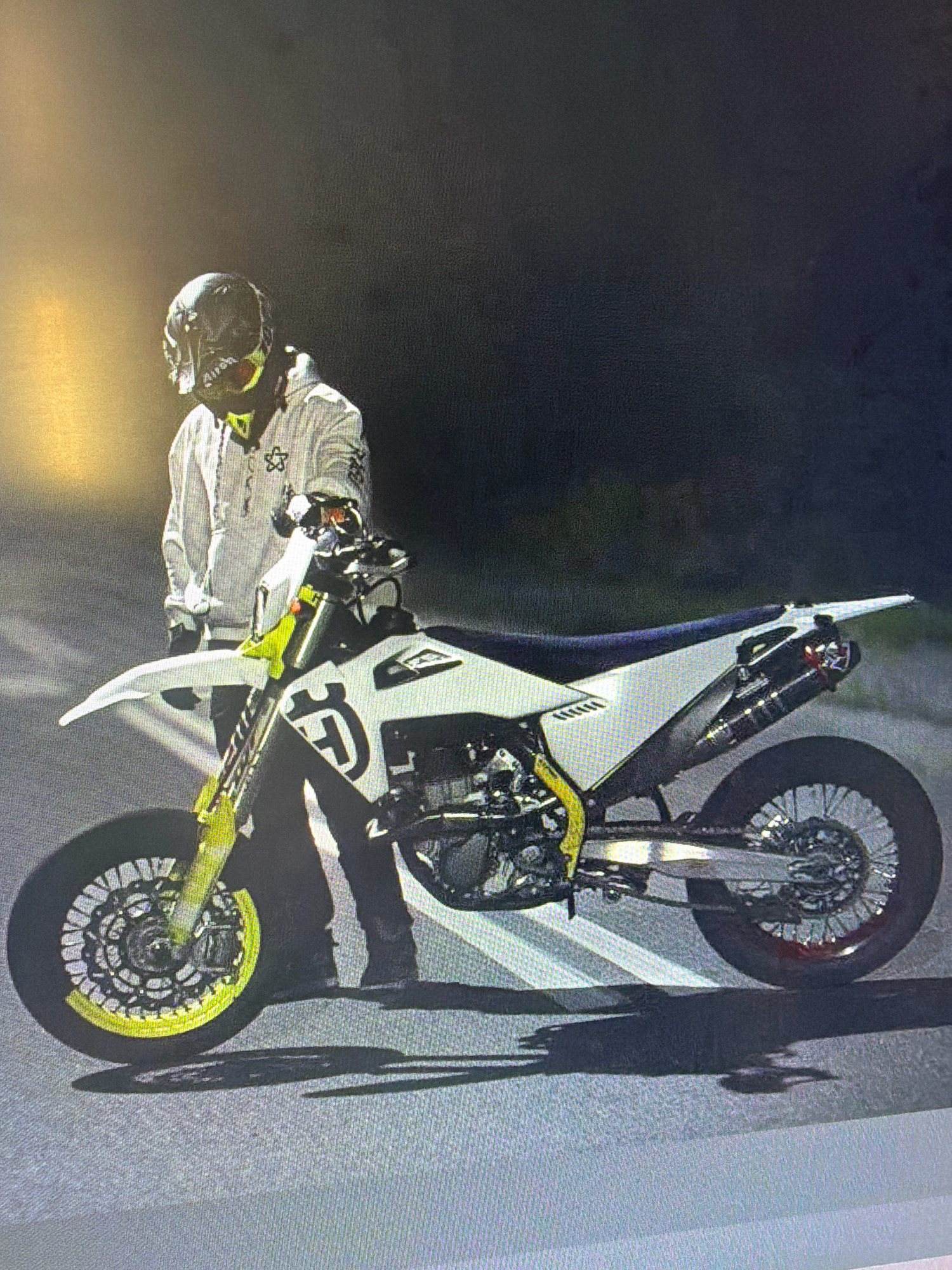 A person wearing a white hoodie and a helmet stands beside a white and black dirt bike. The dirt bike has yellow accents on the wheels and handlebars. The person is holding onto the handlebars of the dirt bike. The scene appears to be outdoors at night, with a dark background and some artificial lighting. The ground is paved, possibly a street or parking lot. The image is a still photograph. There is no visible indication of the location being Newark, United States of America.