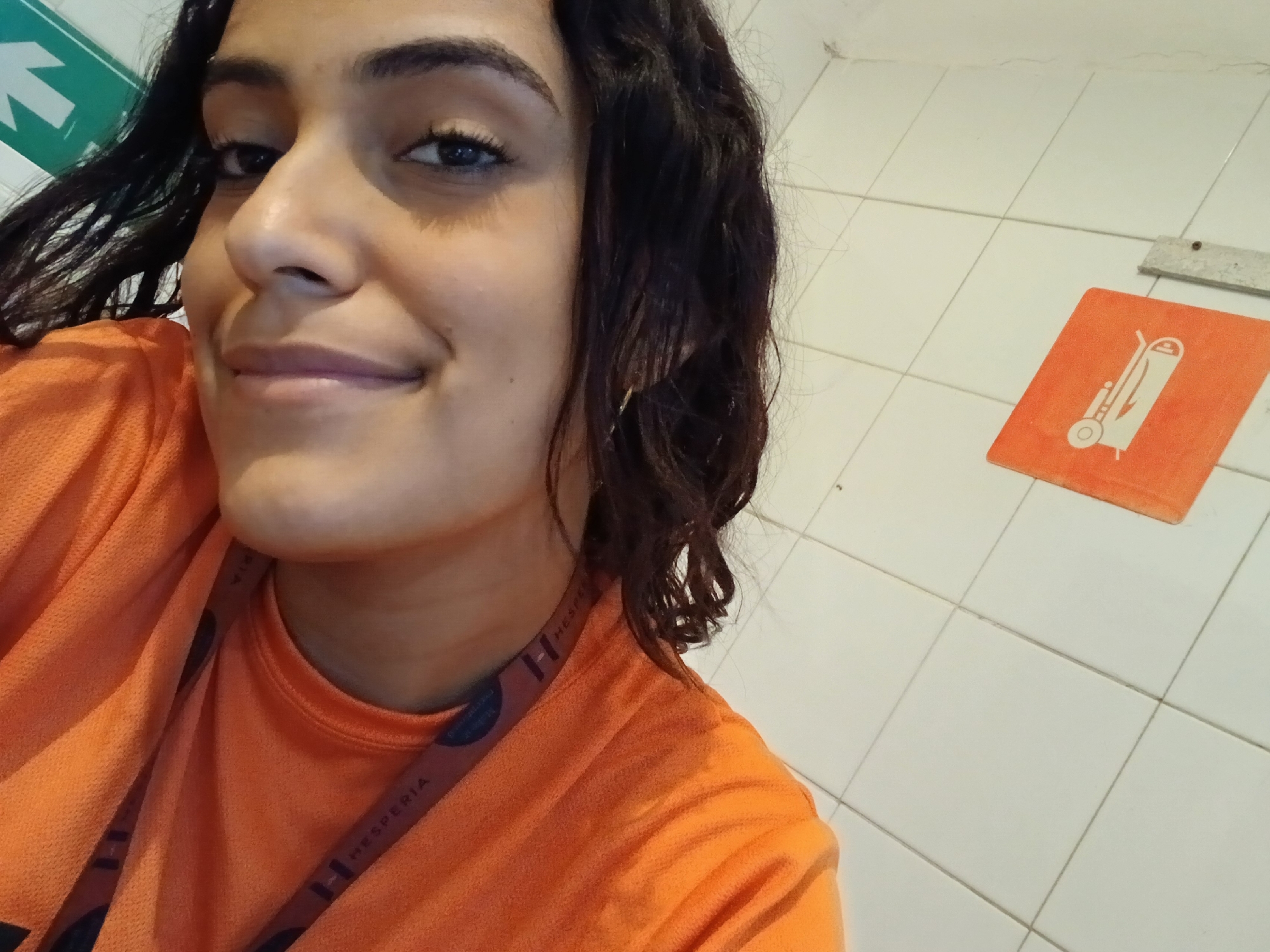 A young woman with dark, wavy hair and a subtle, warm smile is featured in a close-up shot. Her dark eyes are looking towards the camera. She wears a bright orange t-shirt, which appears to be part of a uniform. Around her neck, a lanyard is visible with the text "HESPERIA" printed on it, along with an 'H' logo.
The background consists of a white tiled wall, typical of a service area, corridor, or utility space within a commercial building. In the upper left corner, a green sign with a white arrow pointing upwards and to the right is visible, likely an emergency exit or directional sign. On the right side of the wall, an orange rectangular sign displays a white icon resembling a cleaning cart with a mop bucket and wringer, suggesting a cleaning area or maintenance-related signage.
The woman's gentle smile conveys a friendly and approachable demeanor. The visible "HESPERIA" text on her lanyard, combined with the context of Playa El Agua, Venezuela, strongly suggests the setting is a Hesperia hotel or resort in that location. The image captures a casual moment, possibly a selfie, during her work shift.