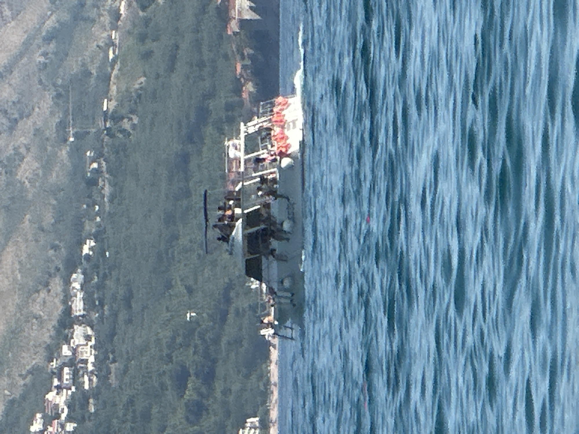 A day time, high-angle, wide shot shows a large, multi-level boat filled with people on a body of water. The boat is white with a gray top level and appears to be a passenger ferry or tour boat. It has orange life preservers visible on the back. The boat is in motion, creating a wake behind it.
In the background, a hillside is covered in green trees. Higher up on the hill, buildings with white walls and red roofs are visible, characteristic of the town of Boreti, Montenegro. The sky is a hazy, light blue. The water in the foreground is a deep blue with visible ripples and waves. There are no apparent interactions between people on the boat or with the viewer.