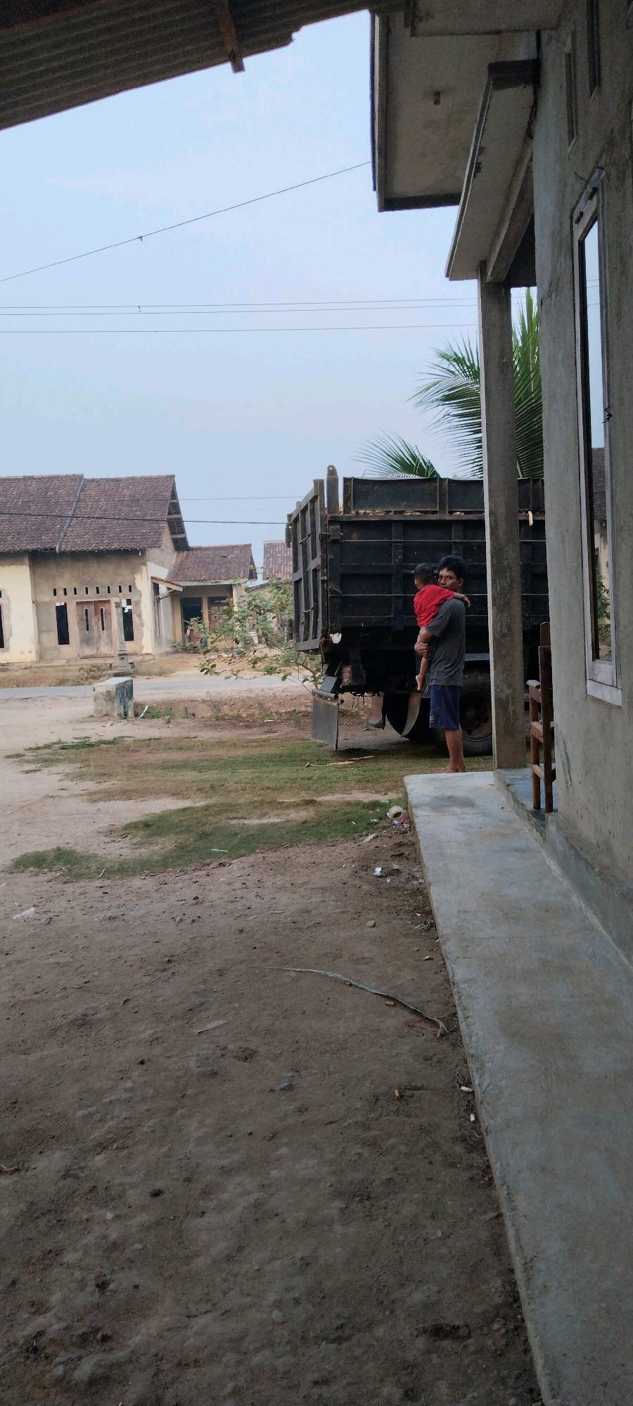 A man in a dark grey t-shirt and blue shorts is holding a young child wearing a red t-shirt. They are standing near the back of a black dump truck. The background shows several houses with tiled roofs and some trees. The foreground is a dirt and grass area. The scene appears to be in a residential area in Tanjung Ratu Ilir, Indonesia.