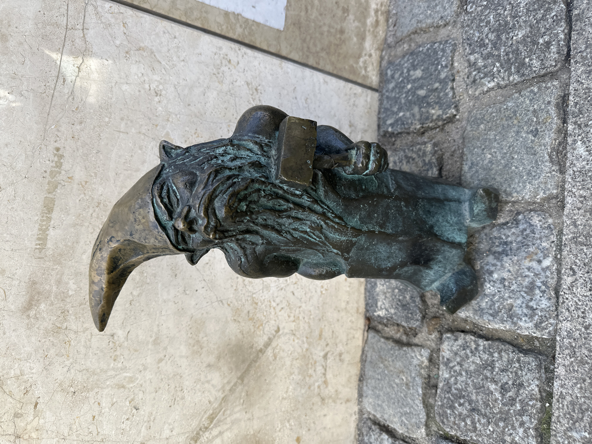 A bronze dwarf statue in Wrocław, Poland, is mounted on the corner of a building. The statue depicts a dwarf with a long, hooked nose, wearing a hat, and holding a hammer. The statue is affixed to a stone wall on one side and a light-colored, marbled wall on the other. The bronze has a weathered, slightly greenish patina.