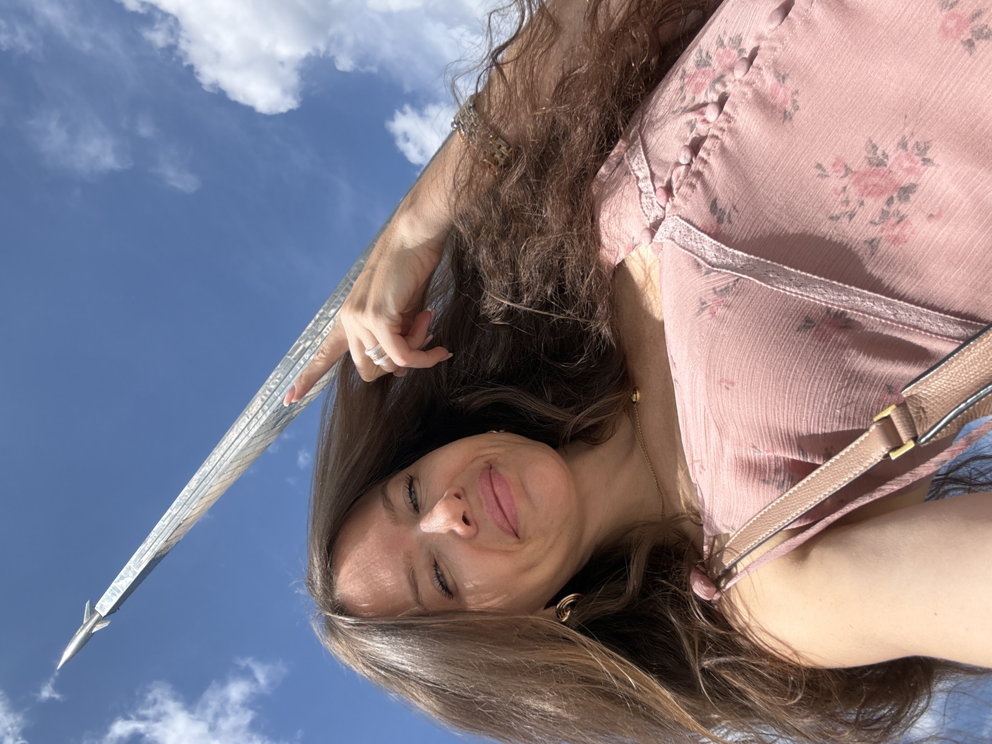 A woman is taking a selfie outdoors on a sunny day. She has long, brown hair and is wearing a light pink, floral-patterned dress and a gold necklace. She is pointing at a tall, metallic monument in the background, which appears to be a rocket or spire, with a small rocket on top. The monument is set against a bright blue sky with white, fluffy clouds. A beige purse strap is visible across her chest. The monument resembles the Monument to the Conquerors of Space in Moscow, Russia.