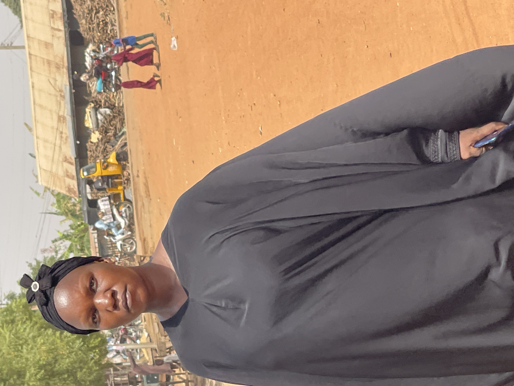In Muchiya, Nigeria, a woman wearing a black hijab stands in an outdoor setting. She faces the camera, holding a phone in her right hand. The background includes a street scene with various motorcycles and a yellow commercial vehicle. Several people are walking in the distance. The ground appears to be reddish-brown, and there are trees and buildings nearby.