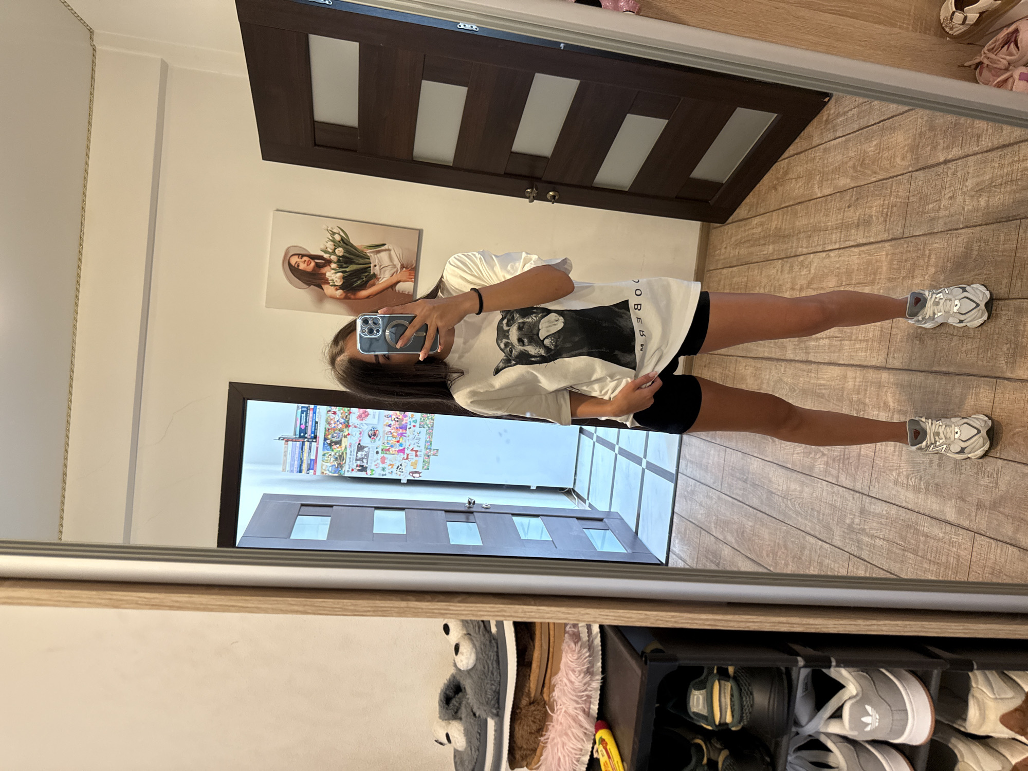 The image shows a young woman taking a mirror selfie in what appears to be a domestic interior, possibly in Chișinău, Moldova. She is standing in front of a large mirror that reflects a portion of a room. She is wearing a white oversized t-shirt with a black dog graphic and black shorts, along with grey and white athletic shoes.
In the reflection, to her left, is a multi-tiered shoe rack filled with various shoes and slippers, including a pair of grey fuzzy slippers with eye details. Beyond the shoe rack, a doorway is visible, leading into another part of the house. The doorway is flanked by a wall adorned with numerous colorful stickers or posters.
To the right of the woman in the reflection, a dark brown wooden door with rectangular glass inlays is prominent. Above this door, a framed picture of a woman wearing a hat and holding a bouquet of white flowers is hung on the wall. The floor is made of light brown wooden planks, and a tiled floor is visible in the background through the open doorway. The overall impression is a casual, personal space, likely a bedroom or hallway in a residence.