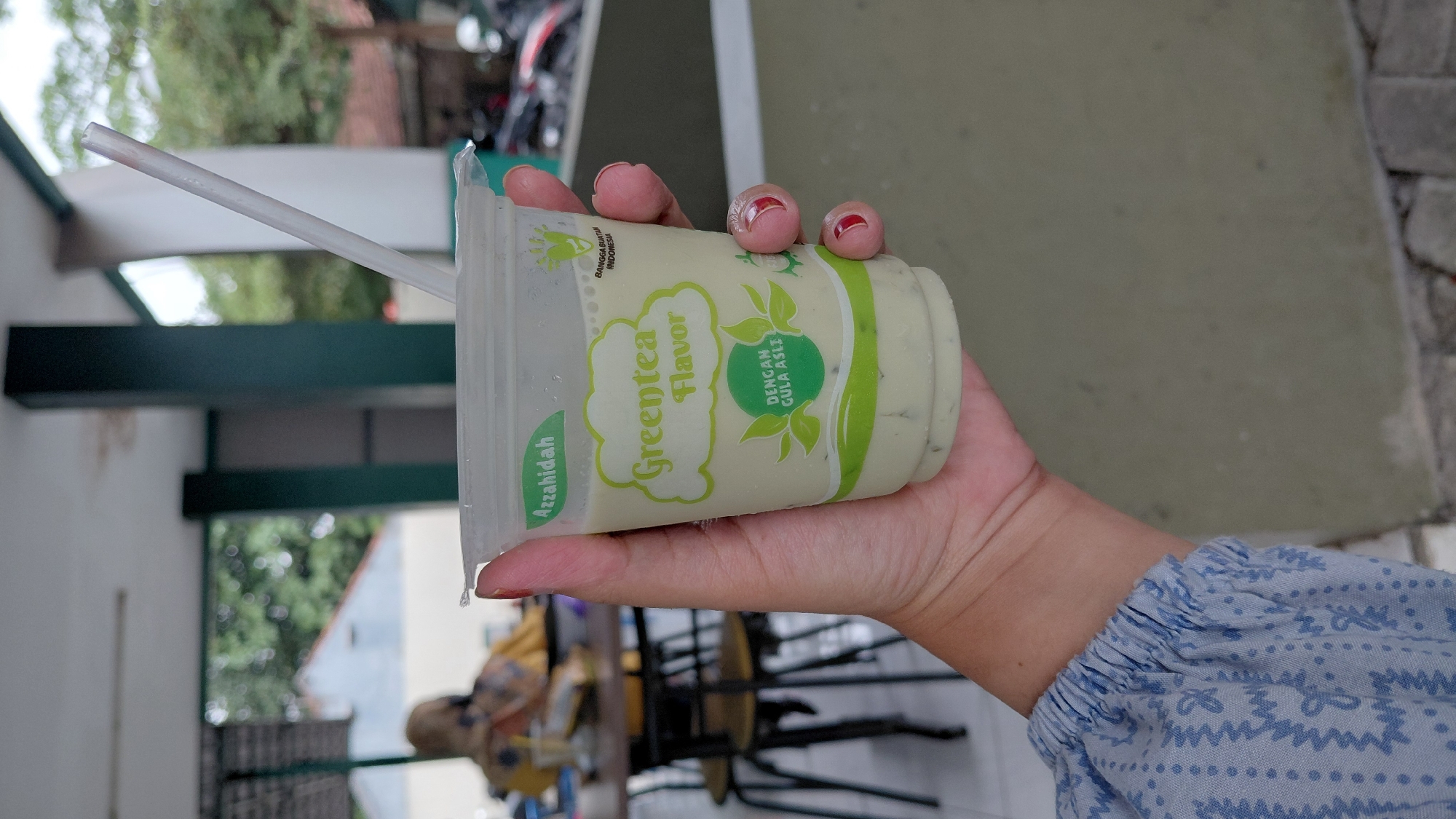 A person's hand is holding a clear plastic cup with a green liquid and a white straw inserted. The cup has a label that reads "Azzahidah Greentea Flavor" with additional text in Indonesian and a green leaf design. The background is out of focus but appears to be an outdoor seating area with tables and chairs, possibly at a cafe or food stall. In the distance, there are buildings and trees. The context suggests this is likely a street-level establishment in Rejasari, Indonesia.