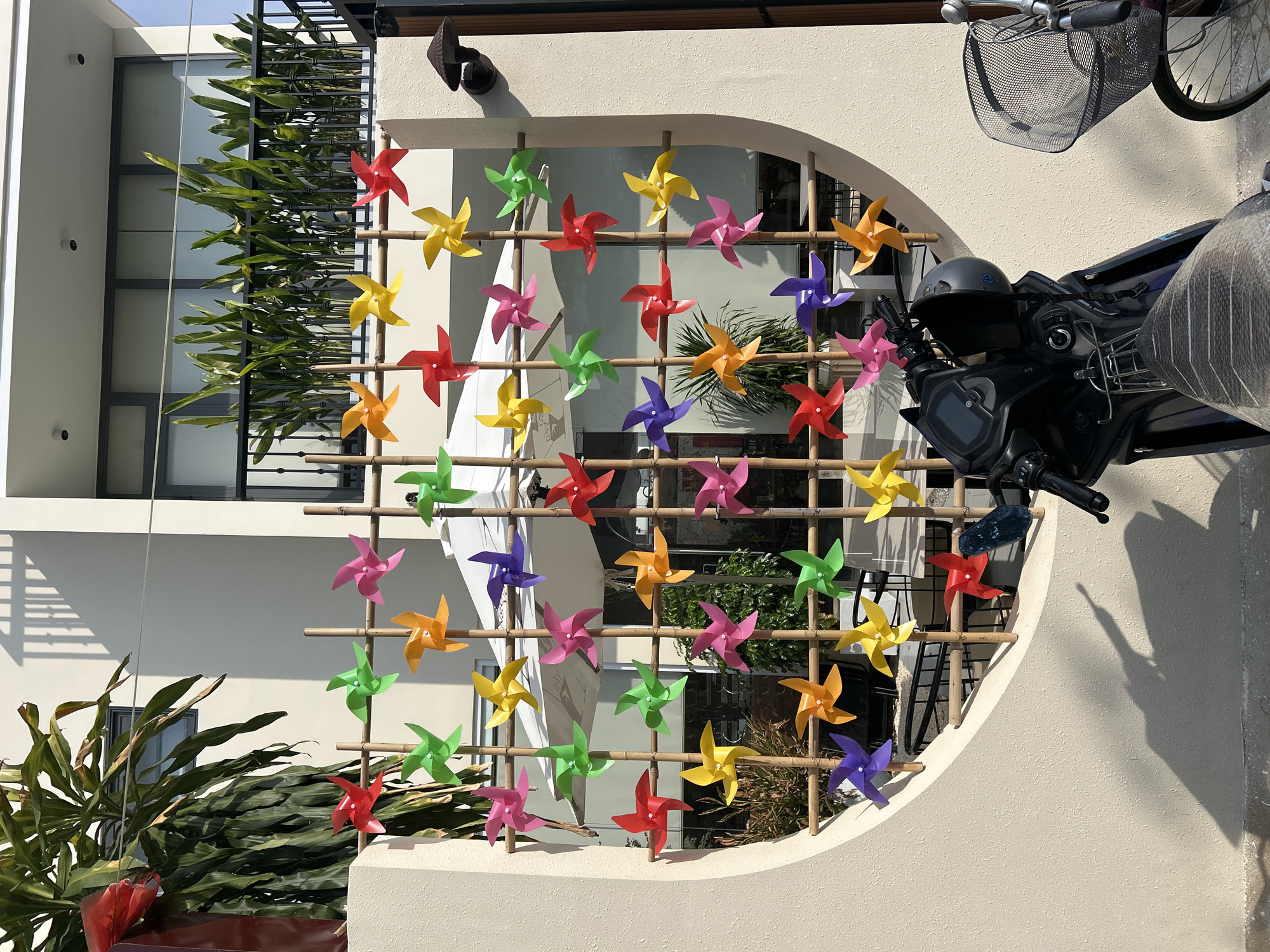 In Nam Nha Trang Ward, Vietnam, a wall decorated with colorful pinwheels made of paper is mounted on a bamboo structure. The pinwheels are red, yellow, orange, green, pink, and purple, and they are arranged in rows. Behind the wall, a modern building with large windows and a balcony is visible. The balcony is filled with lush green plants. In the foreground, a black motorcycle is parked, and a bicycle with a wire basket is also visible. The scene is bathed in sunlight, casting shadows on the wall.