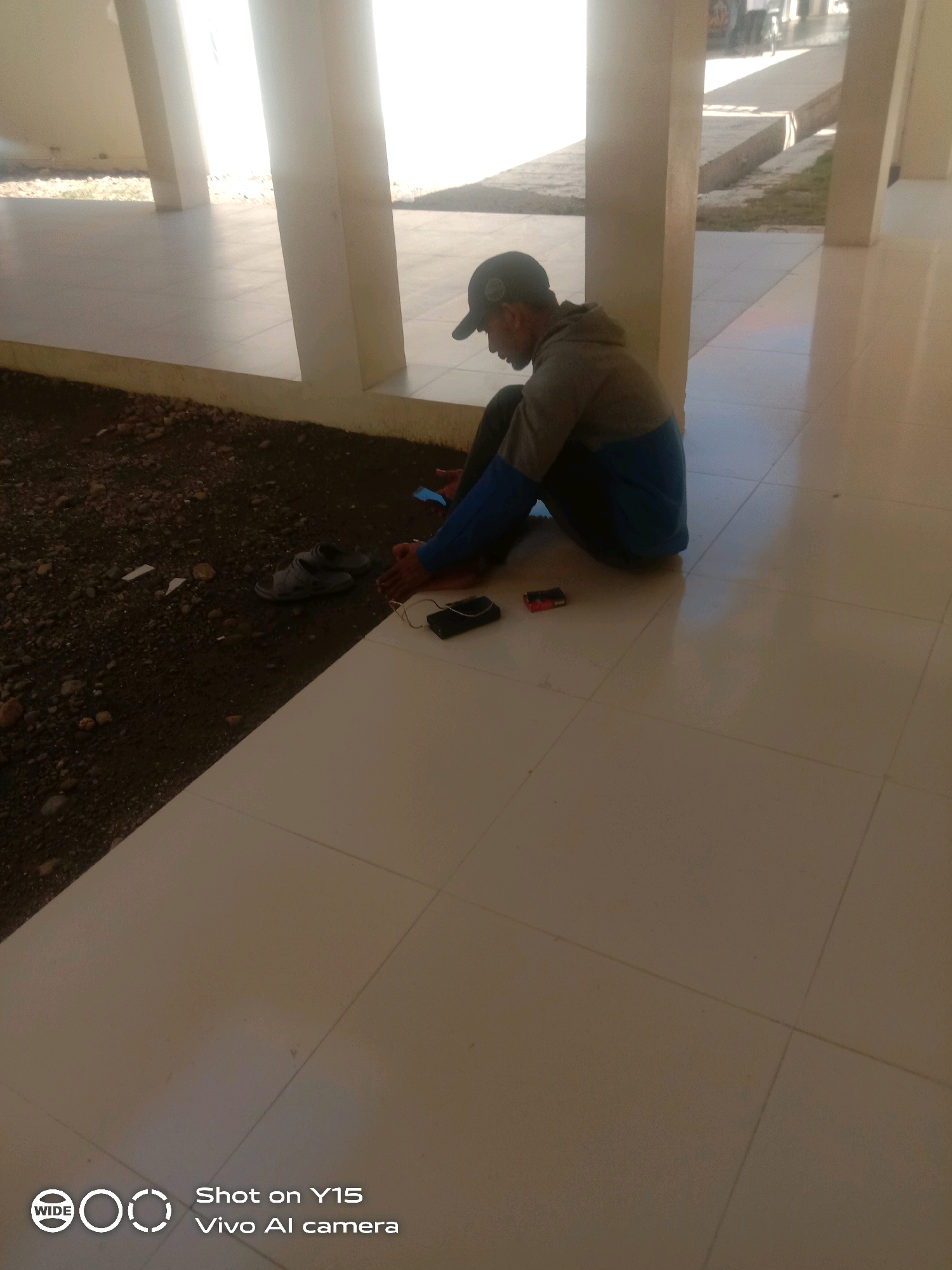 A man in a baseball cap and hooded jacket sits on a tiled floor outside a building, with his back to a patch of dark soil. Two sandals are placed on the soil near him, and a black rectangular object, possibly a phone or charger, lies on the tile in front of him. The man is looking down at his lap. The environment appears to be an open-air covered walkway or patio with white tiled flooring and concrete pillars supporting a roof. The lighting suggests it is daytime. The bottom left corner of the image contains a watermark with "WIDE" and "Shot on Y15 Vivo AI camera."