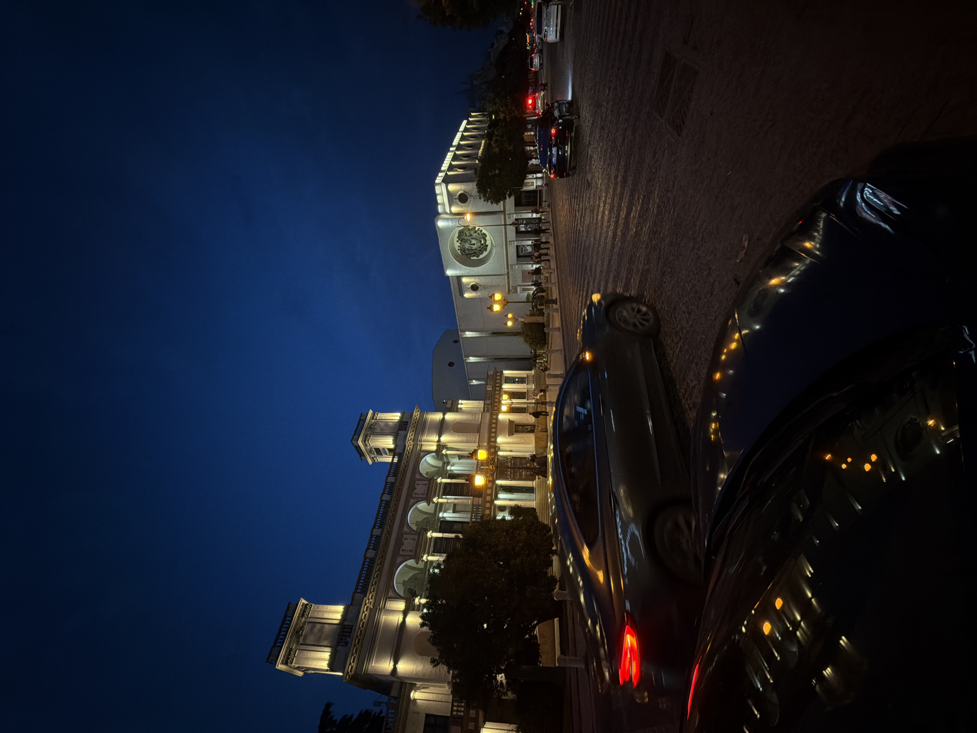 The image depicts a night scene in Tbilisi, Georgia, featuring the illuminated Gabriadze Theater and Rezo Gabriadze Clock Tower. The view is somewhat obscured by parked cars in the foreground. The cobblestone street leads towards the theater. Streetlights and building illumination create a warm glow against the deep blue of the evening sky. Cars are seen on the road near the theater.