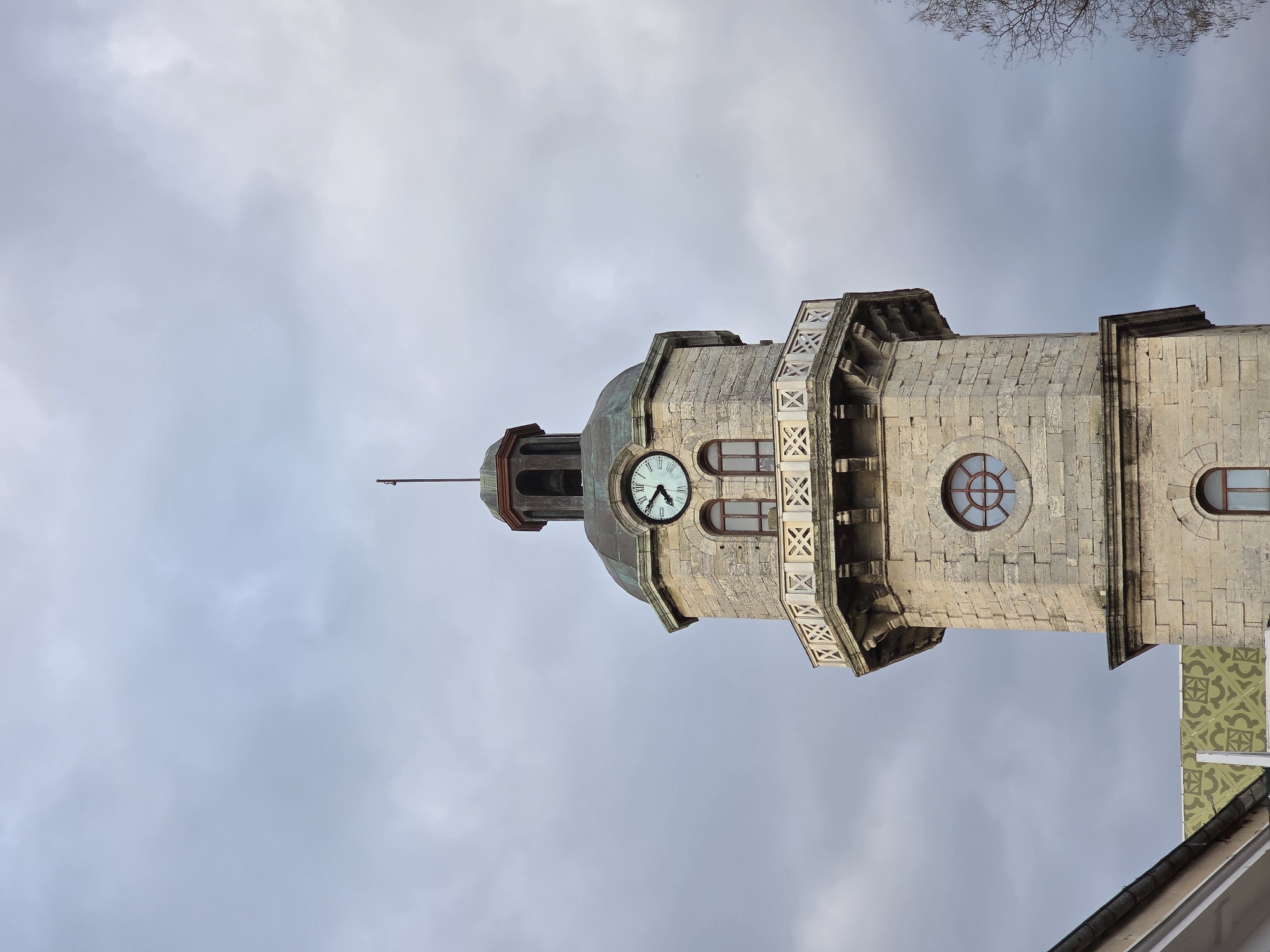 The image depicts the Clock Tower of Varna, Bulgaria, set against a cloudy sky. The tower is constructed from light-colored stone and features a clock face, round windows, and decorative architectural elements. Part of a building roof can be seen at the bottom of the picture. There are bare tree branches at the very top of the image. The tower is tall and narrow, dominating the composition.