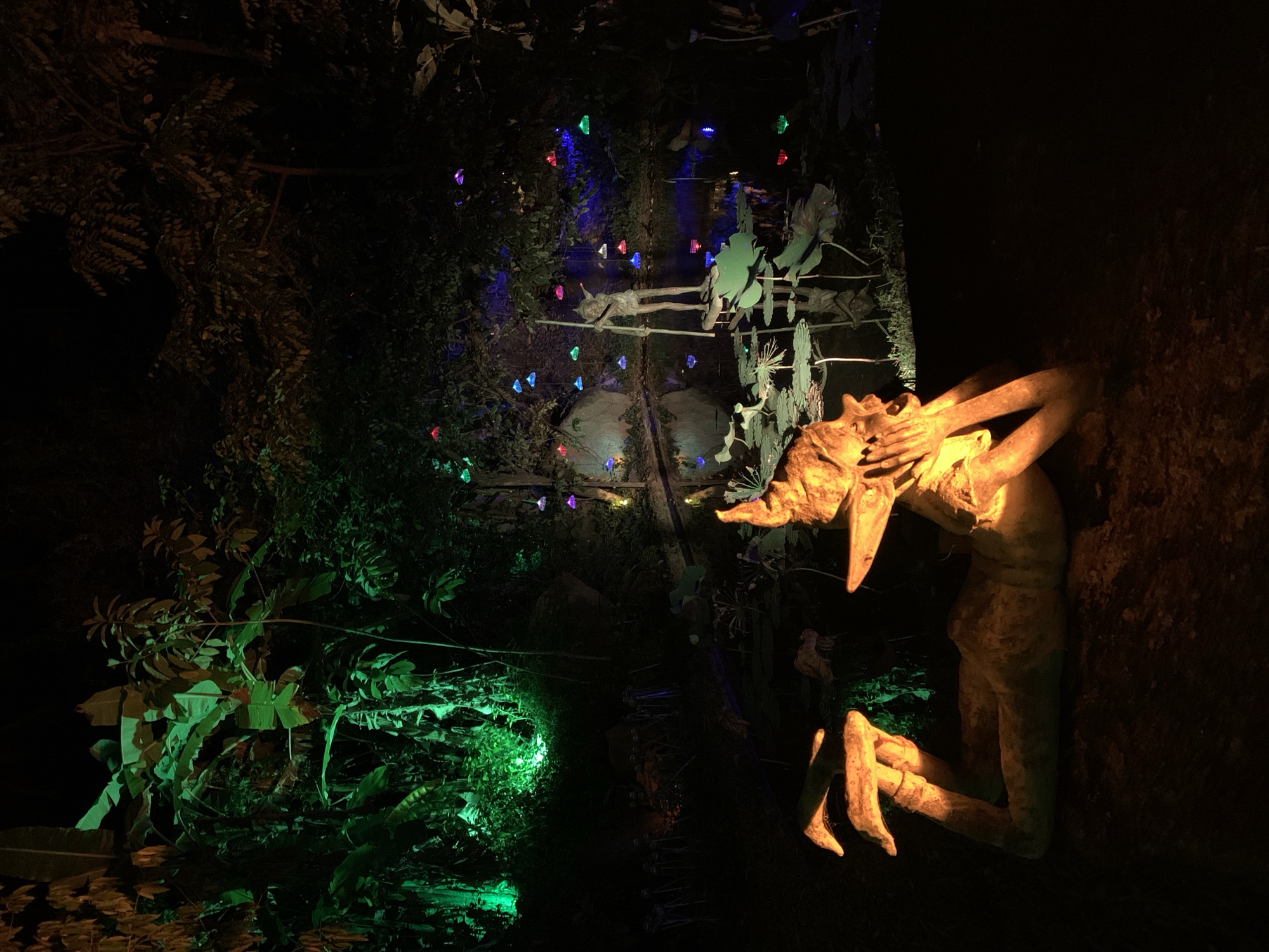 The image shows a nighttime scene in what appears to be a garden or park in Baan Lipa Noi, Thailand. On the right, a statue of a humanoid figure with pointy ears and long fingers is illuminated by a warm light, creating a stark contrast against the dark background. The figure's hands are positioned near its face, suggesting a posture of contemplation or surprise. To the left and further back, there is lush green vegetation, lit by a green light source. In the distance, decorative lights of various colors (blue, red, green) are strung through the trees, adding a festive ambiance. Between the statue and the vegetation, there is a glimpse of a decorative structure with silhouette figures on it, also illuminated by the colored lights. The overall impression is that of an enchanted garden or a display with an element of fantasy.