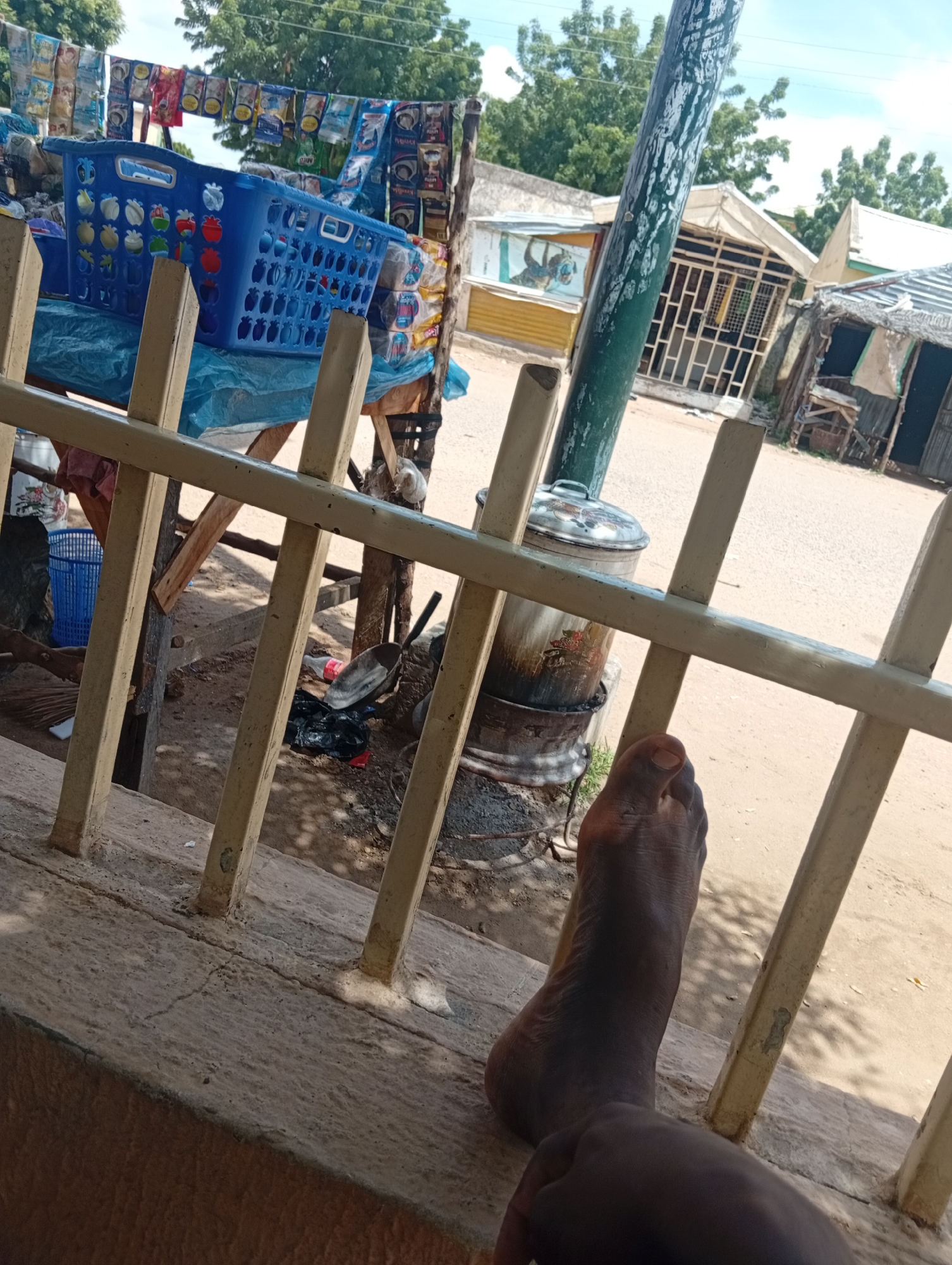 A person's bare foot rests on a concrete ledge in the foreground, casting a shadow. Behind the foot, a light-colored metal railing divides the frame. In the middle ground, a blue plastic basket sits on a table covered with blue plastic sheeting, surrounded by a variety of packaged goods hanging from a line. Beyond this, several small market stalls with corrugated metal and thatched roofs are visible, interspersed with trees and a clear sky. The ground appears to be dirt or sand. The location is identified as the market in Zongoma Kyari, Nigeria.