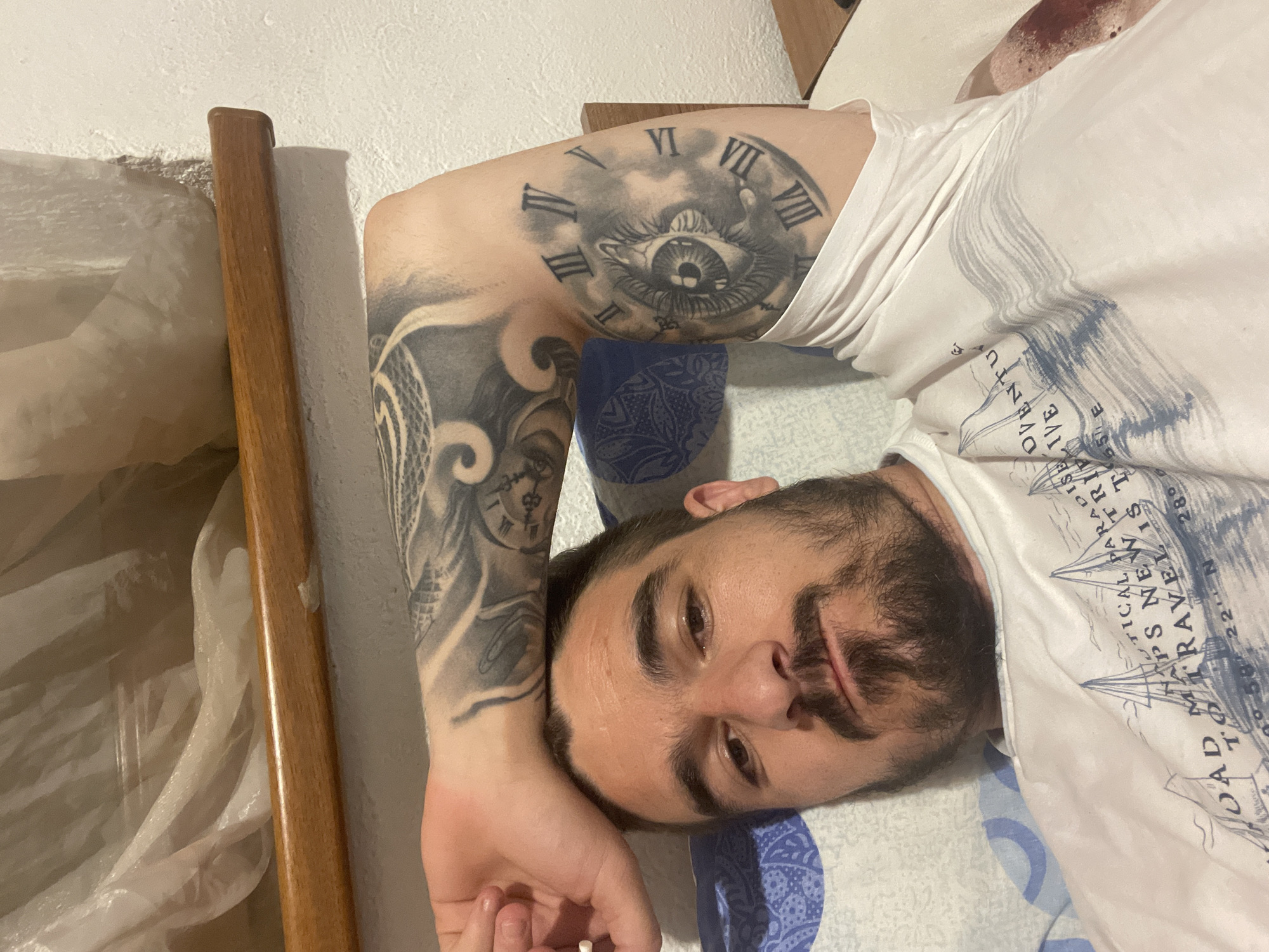 A man with a full beard and mustache is depicted lying down, looking directly at the camera. He is wearing a white t-shirt with a blue graphic print. His left arm is extensively tattooed with black and grey designs. A prominent tattoo on his upper arm features a detailed eye surrounded by Roman numerals (VI, VII, VIII, III, IV, V) resembling a clock face, set within cloud-like imagery. Additional intricate patterns are visible on his forearm. His head rests on a blue and white patterned pillow. In the background, a wooden element, possibly part of a bed frame or headboard, is visible next to a textured white wall.
The white t-shirt features text and nautical-themed illustrations, including sailboats and compass rose elements. Visible text fragments from the shirt include: "OVENTU", "TICAL PARADISE IVE", "HAPS NEW", "TRIP", "JOAD TRAVEL IS T", "T", and coordinates "22 N 28°", suggesting themes of travel and adventure. The man's expression is neutral. The scene appears to be indoors, under artificial lighting.