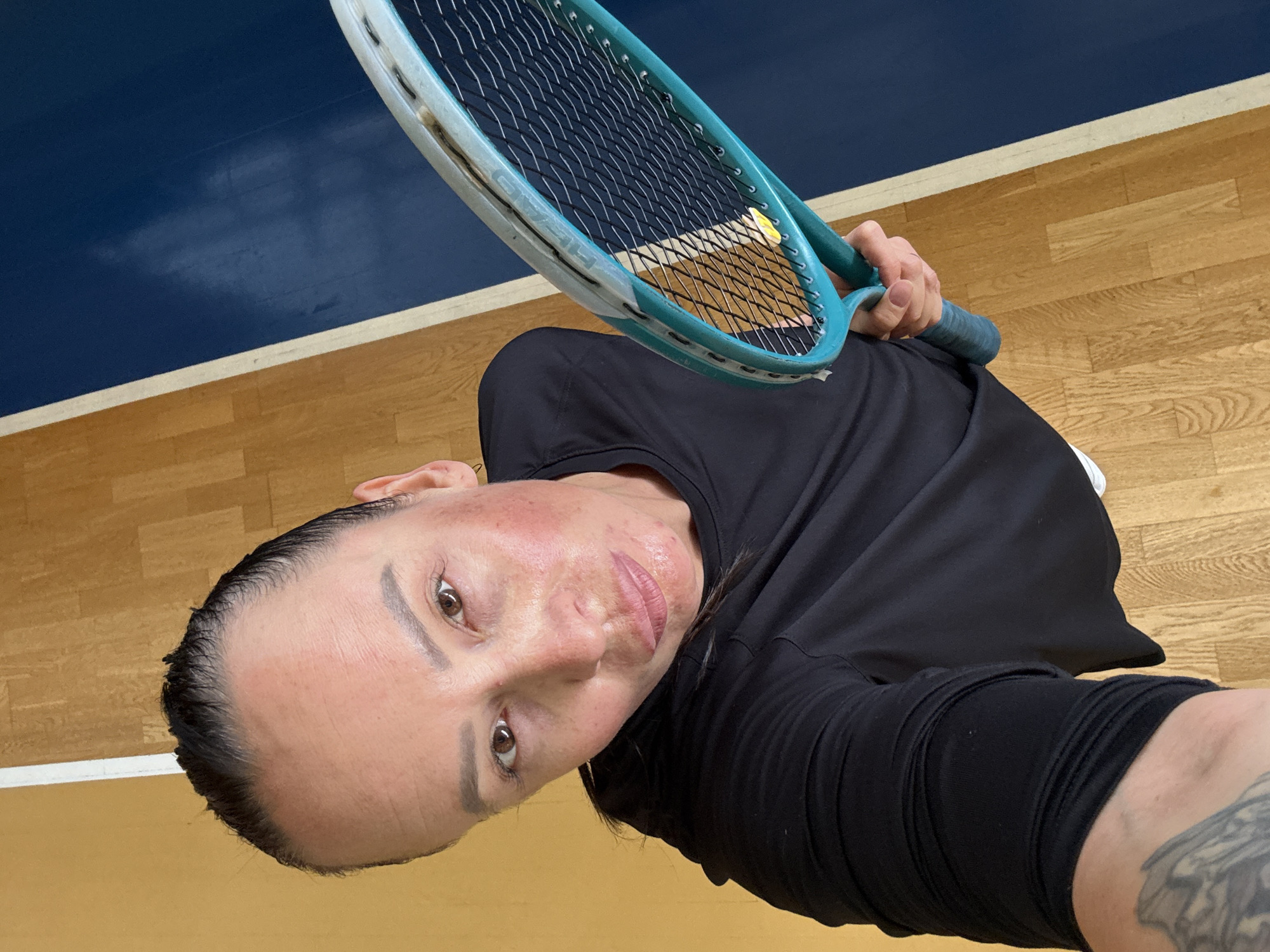 A selfie of a woman holding a tennis racket. She is wearing a black t-shirt and appears to be on a court with a wooden floor and a blue surface. The image is taken from a high angle, looking down at the woman. The location is stated to be in Chaikovsky, Russia.