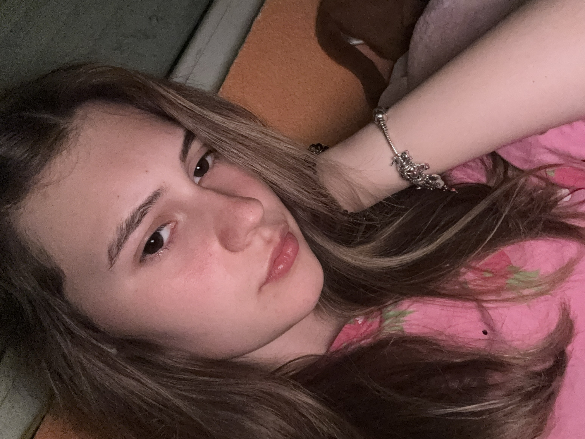 A young woman is lying down, partially angled towards the camera. Her hair is light brown with lighter highlights. She is wearing a pink top. She has a silver charm bracelet on her arm. The background includes a green and orange surface. The location is unknown and there is no indication of the city of Panagyurishte, Bulgaria.